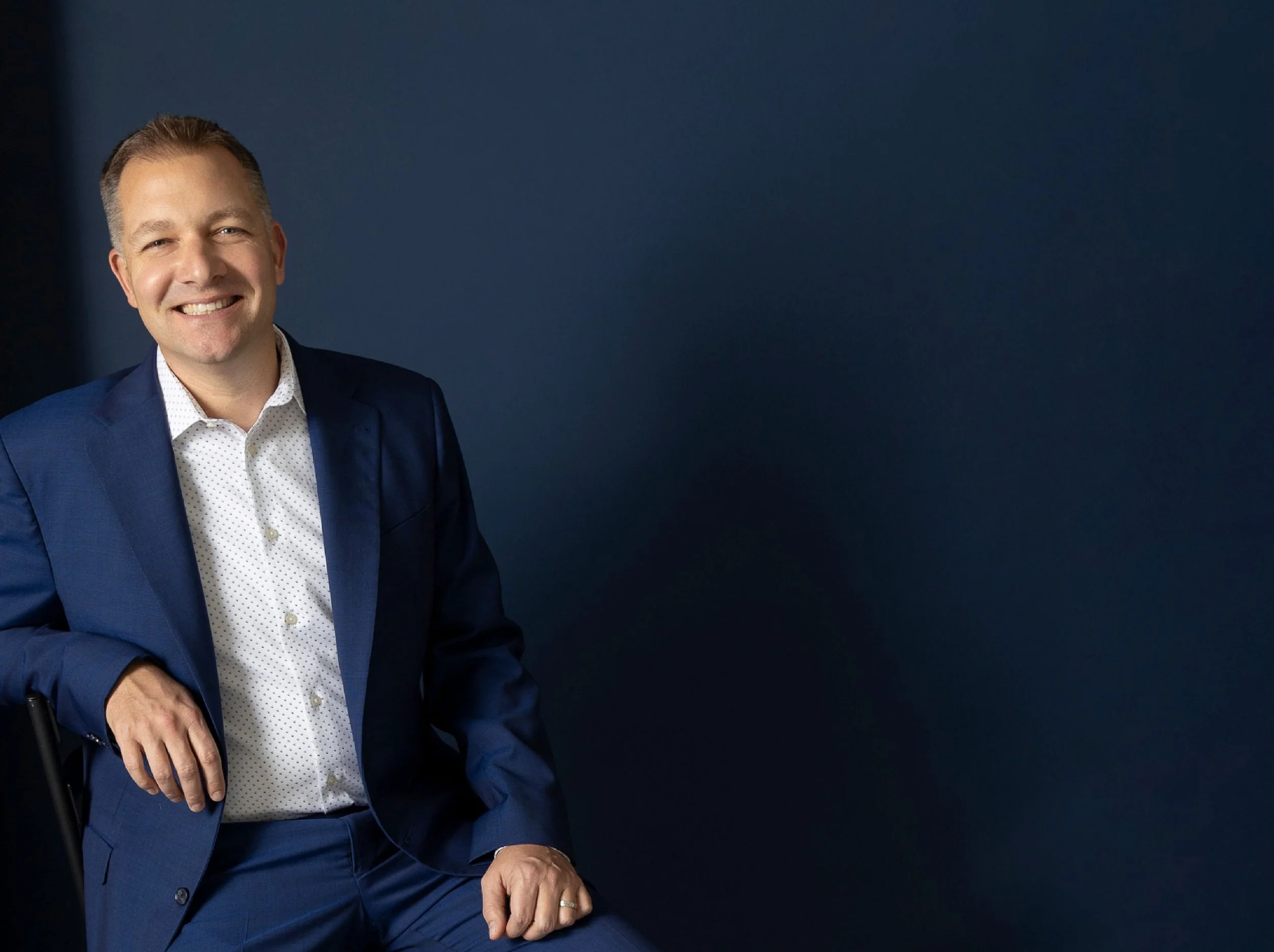 Smiling man in a blue suit sitting on a chair against a dark background.
