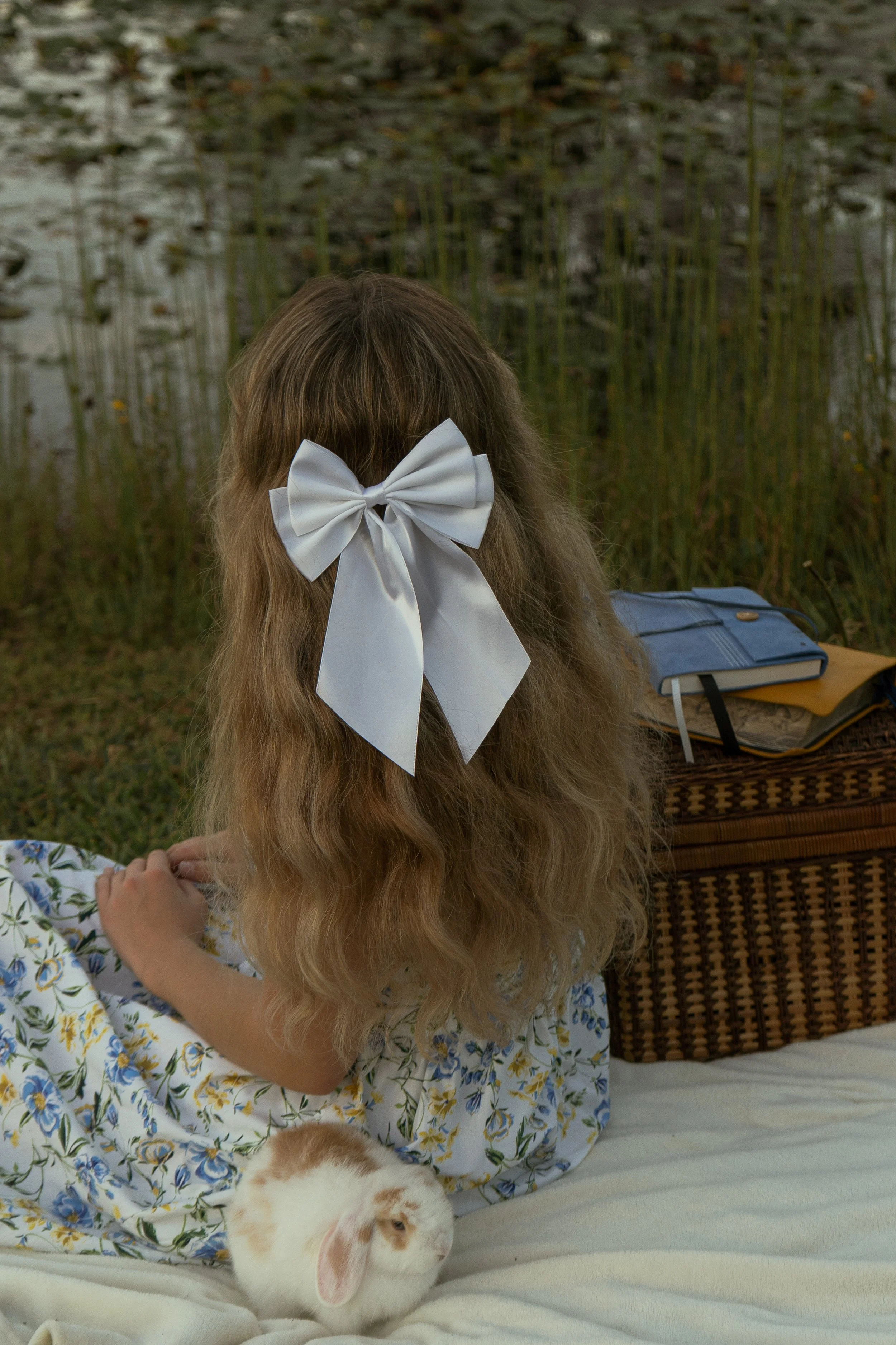 A girl with long hair and a white bow sits on a bed with a rabbit. The girl is wearing a floral dress and appears to be relaxing outdoors by a pond with tall grass and water lilies.