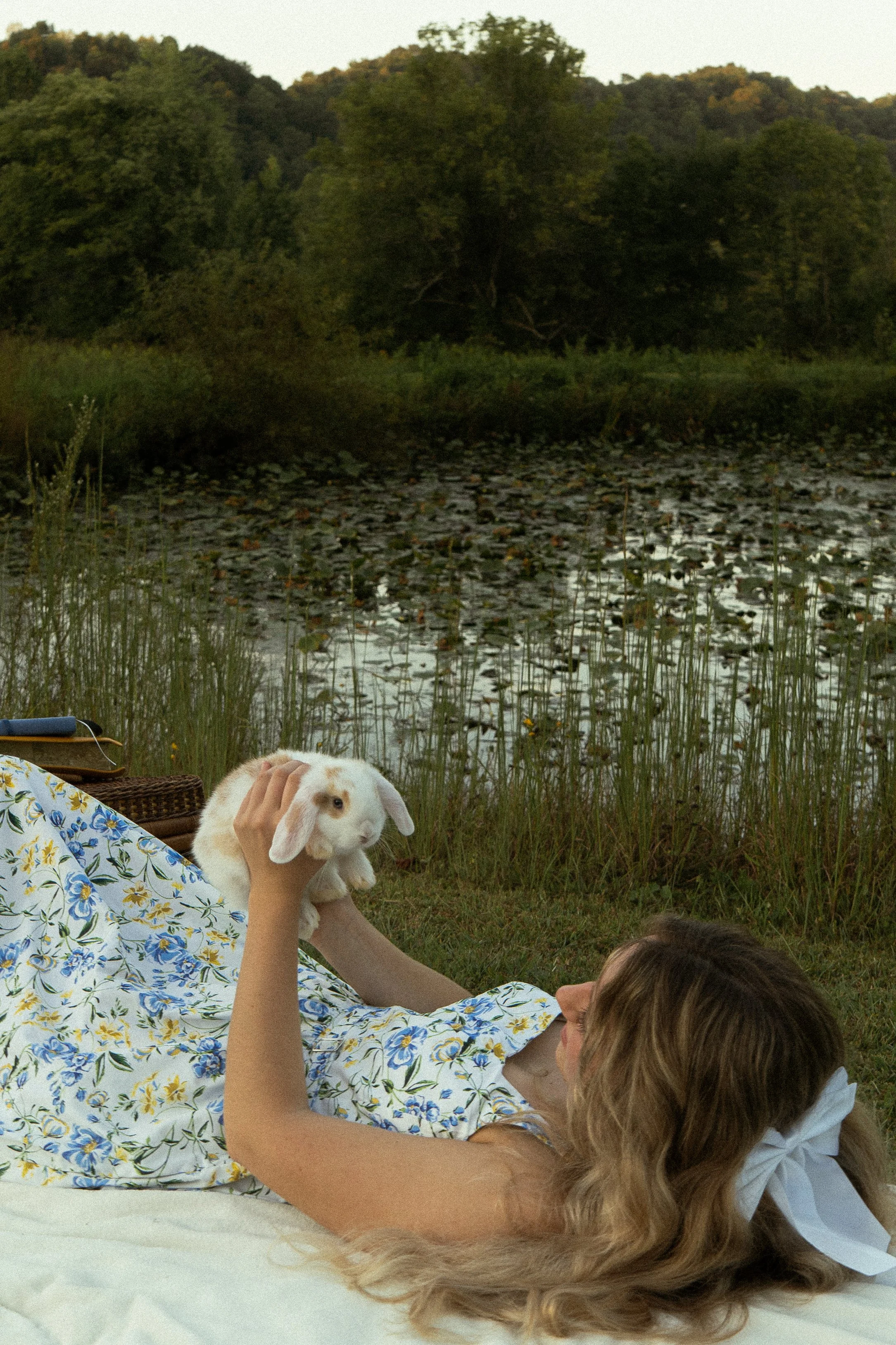 A woman lying on the grass by a lake, petting a white rabbit with brown patches. The woman is wearing a dress with blue and yellow floral patterns. A person with long hair is lying nearby, and a rabbit is also in the scene, with trees and water in th