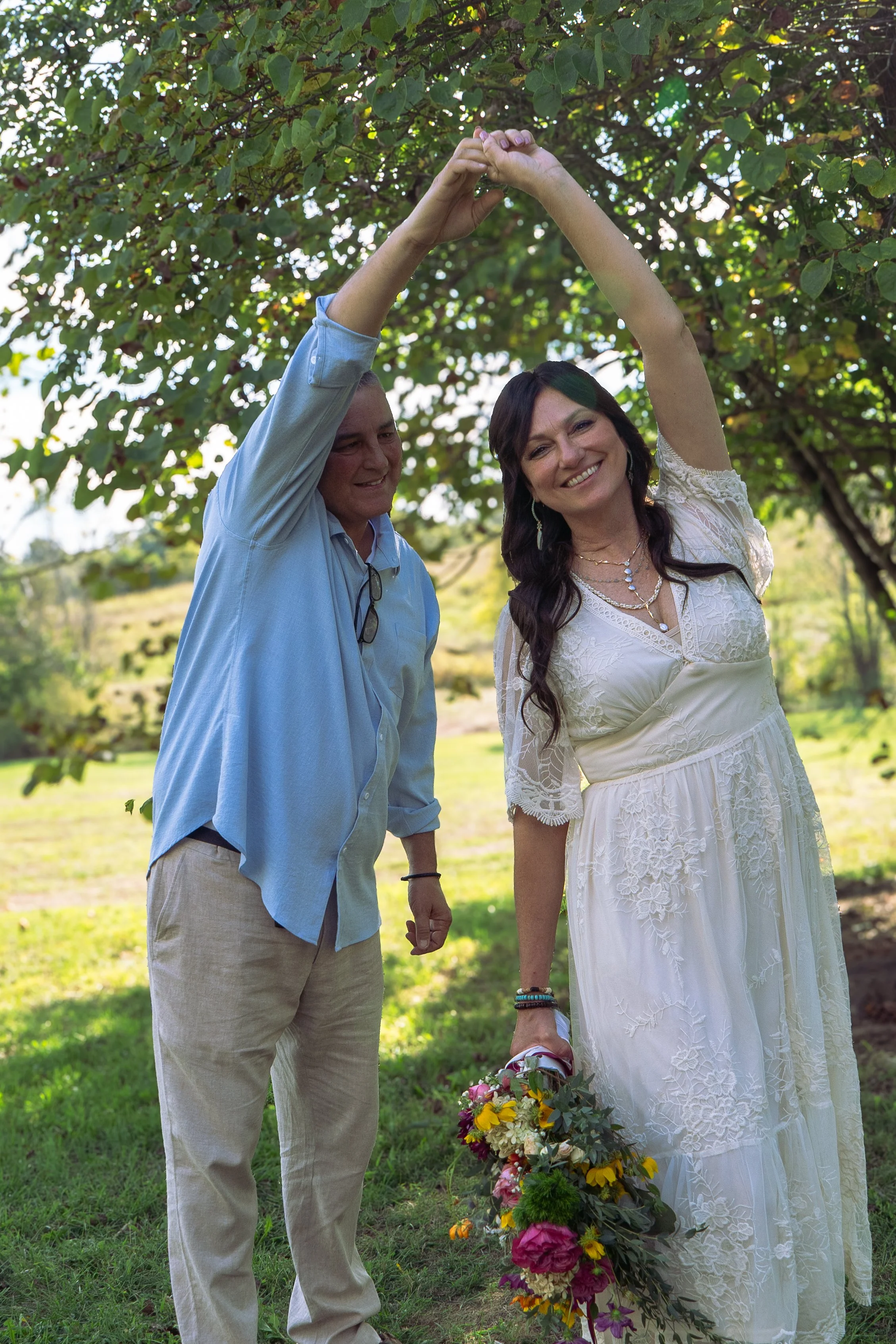 A woman in a white dress is holding a bouquet of flowers at her feet, smiling, while a man in a light blue shirt and beige pants stands next to her, both with their hands joined above their heads as if forming a heart shape, outdoors under green leaf