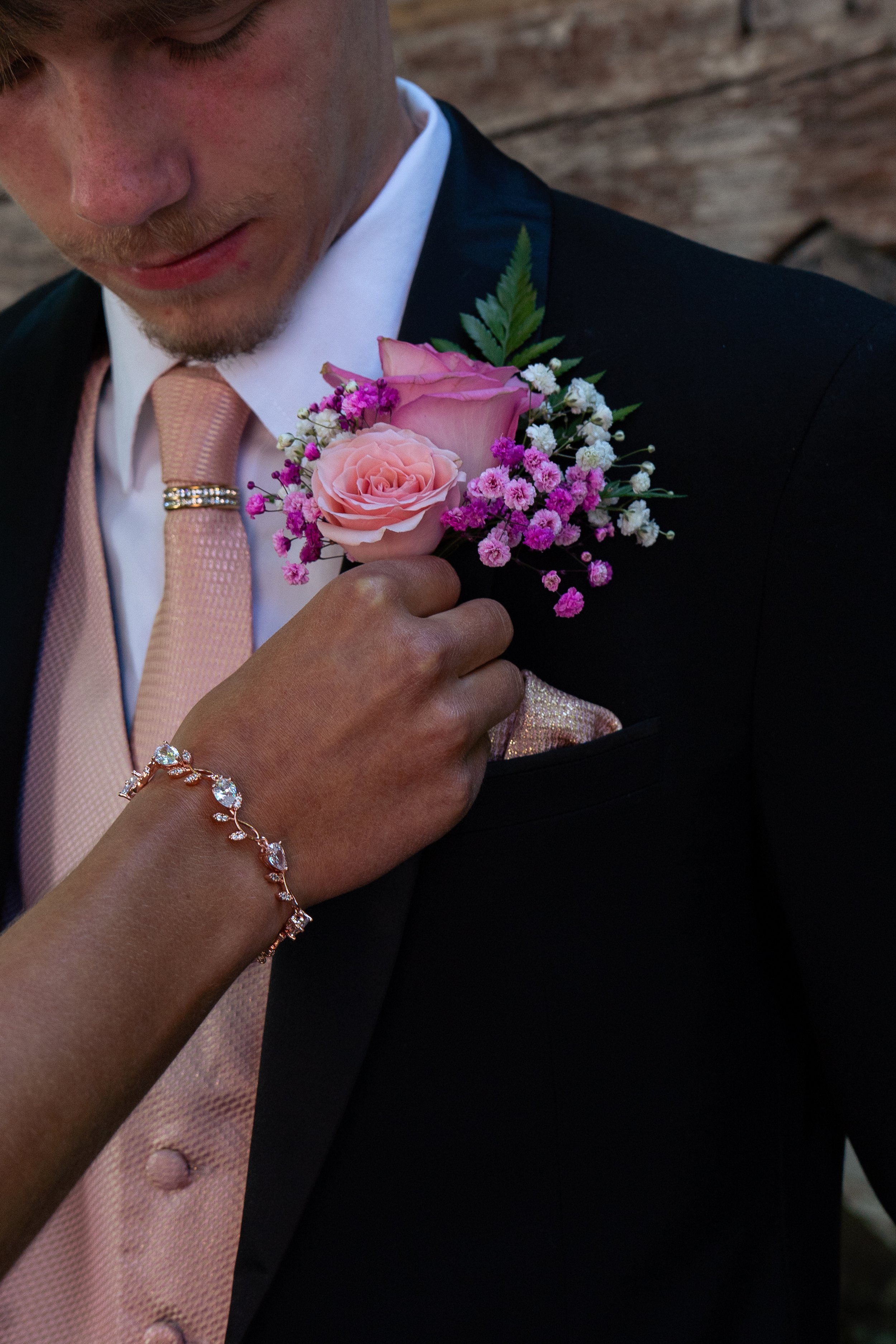 A man in formal dress has a light pink flower boutonniere on his lapel, including pink roses, white and purple small flowers, and green leaves.