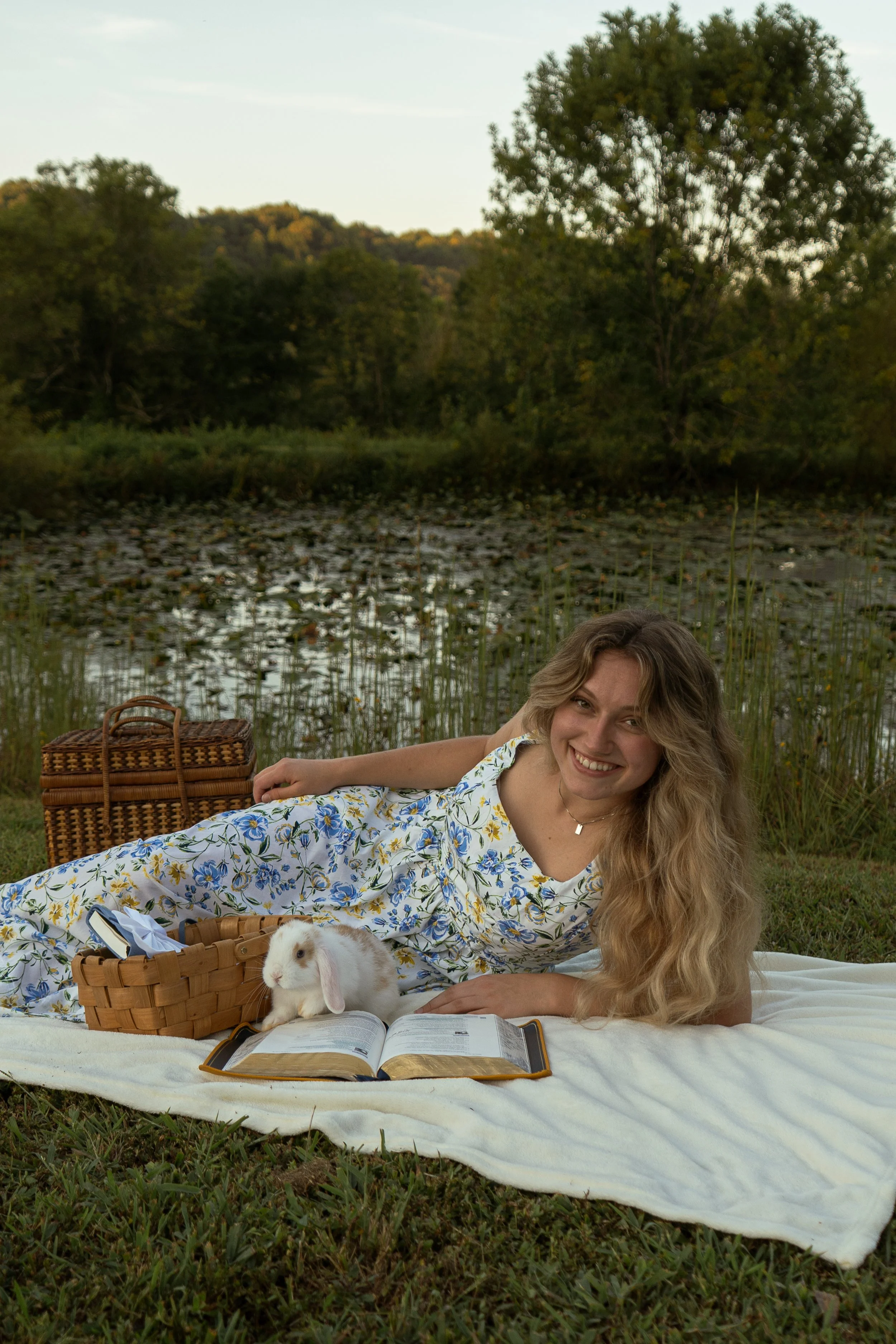 A woman with long, wavy blonde hair lying on a blanket near a pond with lily pads, smiling at the camera. She is dressed in a floral dress, and there is a rabbit sitting next to her with a picnic basket and an open book in front. A wicker picnic bask