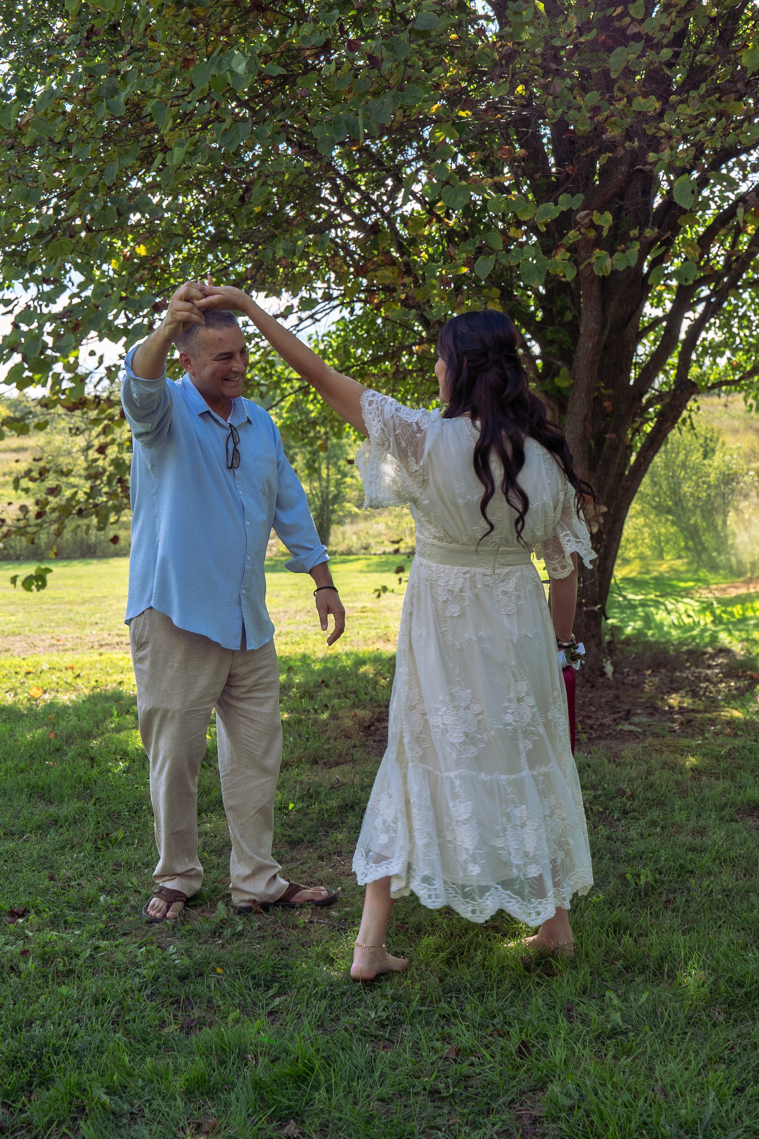 A couple is dancing outdoors under a large tree on a sunny day. The man is wearing a light blue shirt and beige pants, smiling, while the woman, in a white lace dress, has dark hair and is barefoot. They are holding hands.