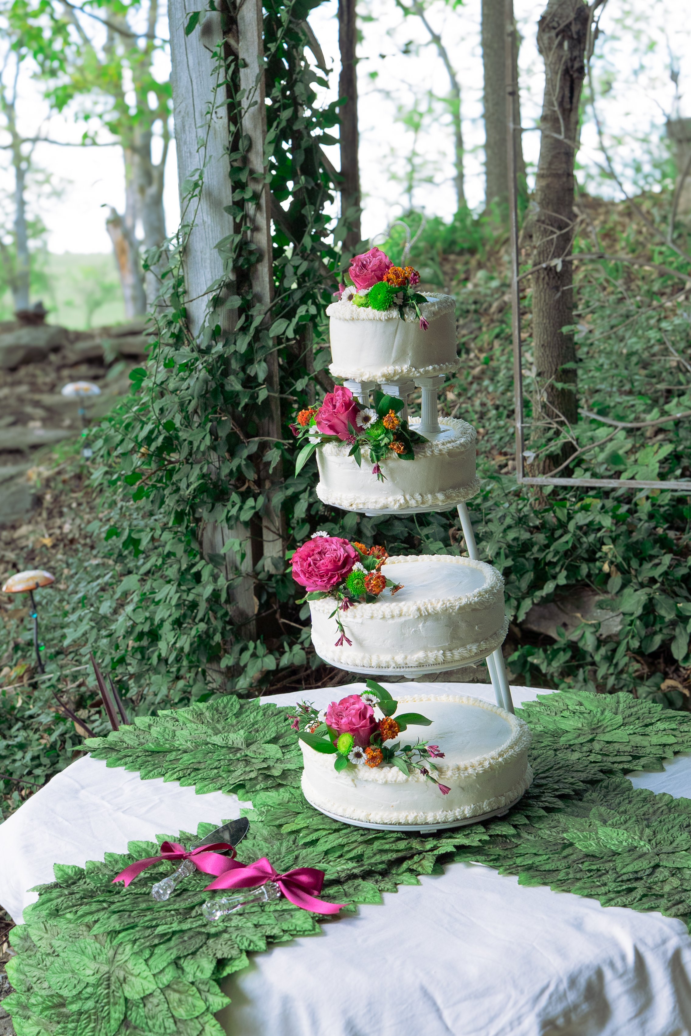 Multi-tiered white wedding cake decorated with pink, green, and orange flowers, placed on a white table with green leaf decorations and a pink ribbon, outdoors in a wooded garden setting.