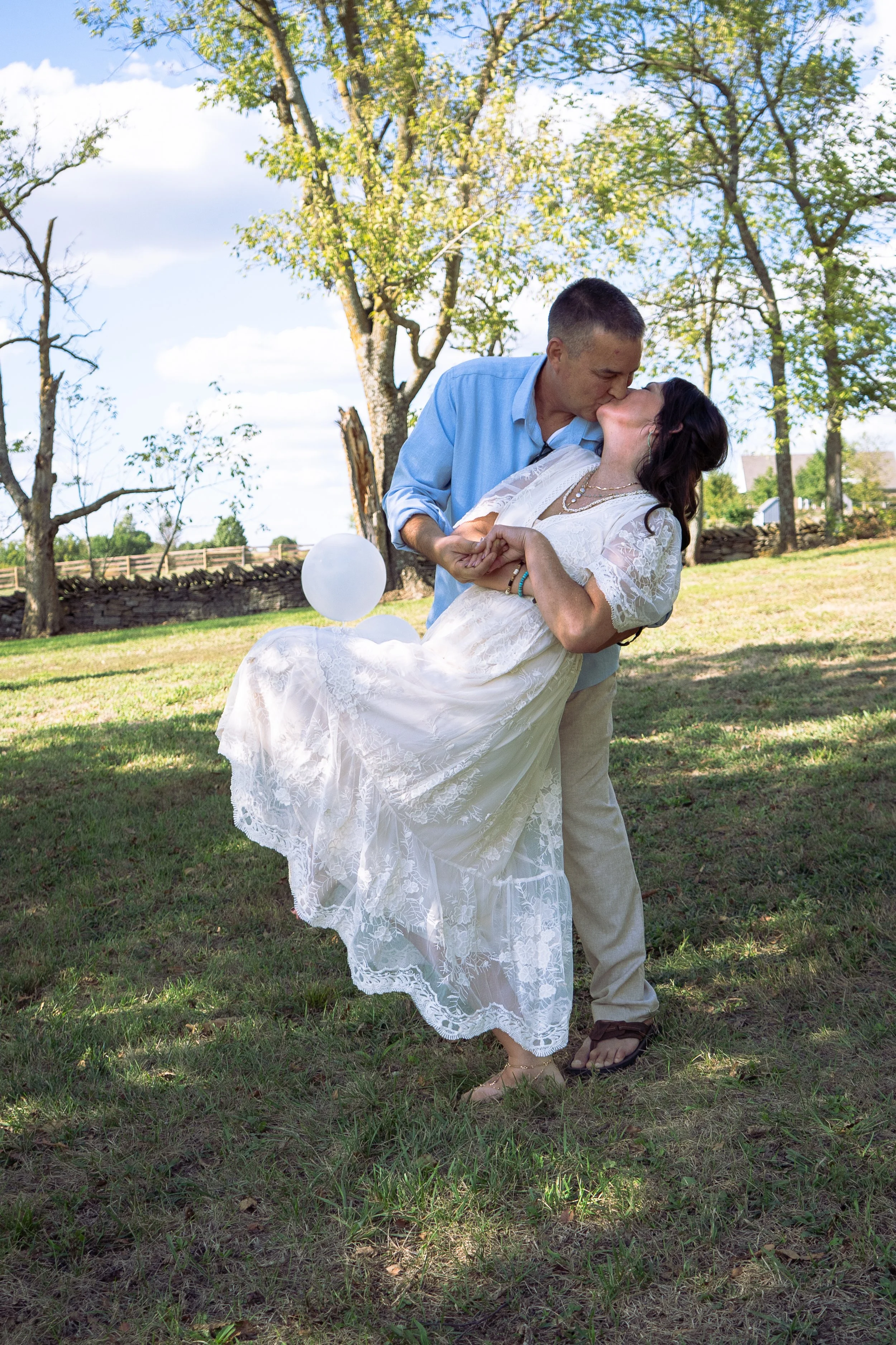 A man in a light blue shirt and a woman in a white lace dress are dancing outdoors on a grassy area with trees and a stone wall in the background. They are sharing a kiss, and the woman is lifting her dress slightly, revealing her feet and sandals. T