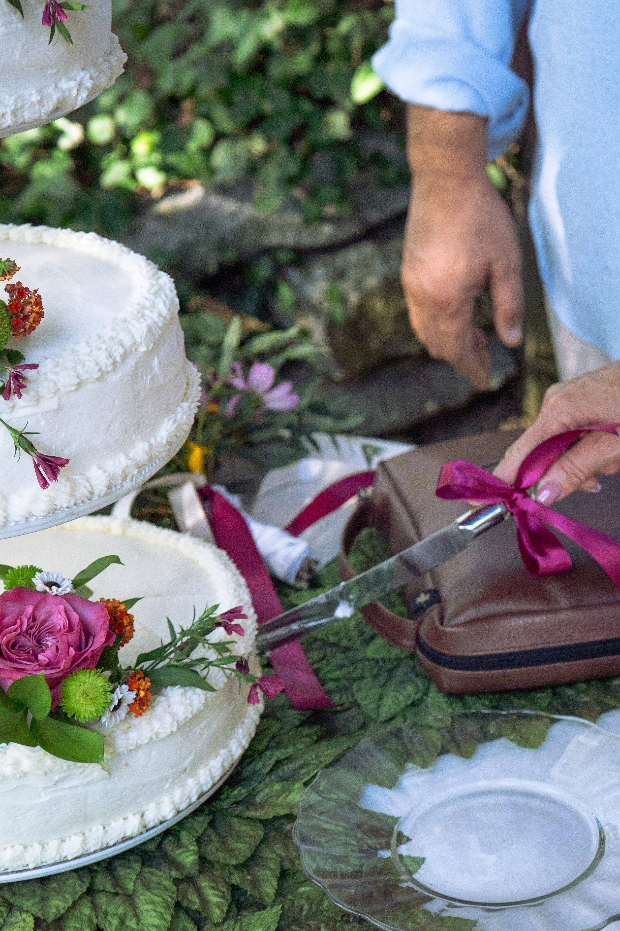 Close-up of a wedding cake decorated with white icing and colorful flowers, with a person slicing it using a knife tied with a pink ribbon, set outdoors on a leafy surface.