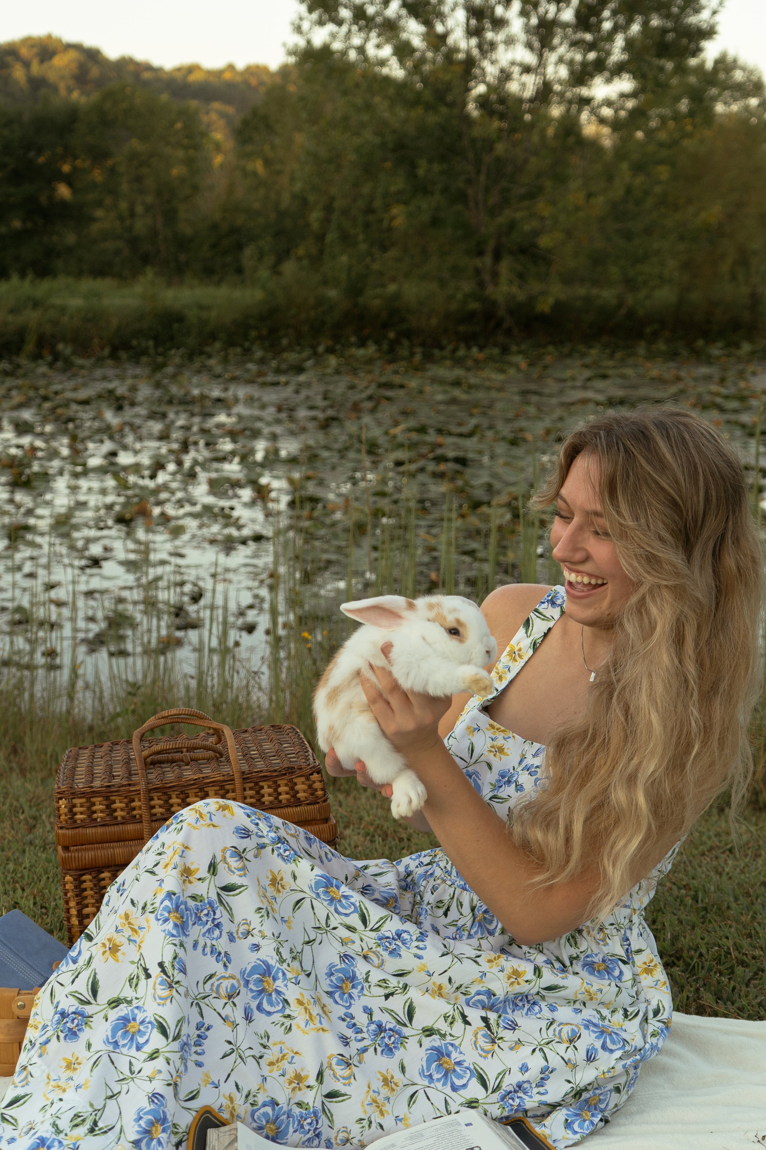 A woman with long, wavy blonde hair sitting outdoors by a pond with water lilies, smiling as she holds a fluffy white and brown bunny. She is wearing a white dress with a blue and yellow floral pattern, with a wicker picnic basket nearby. The scene i