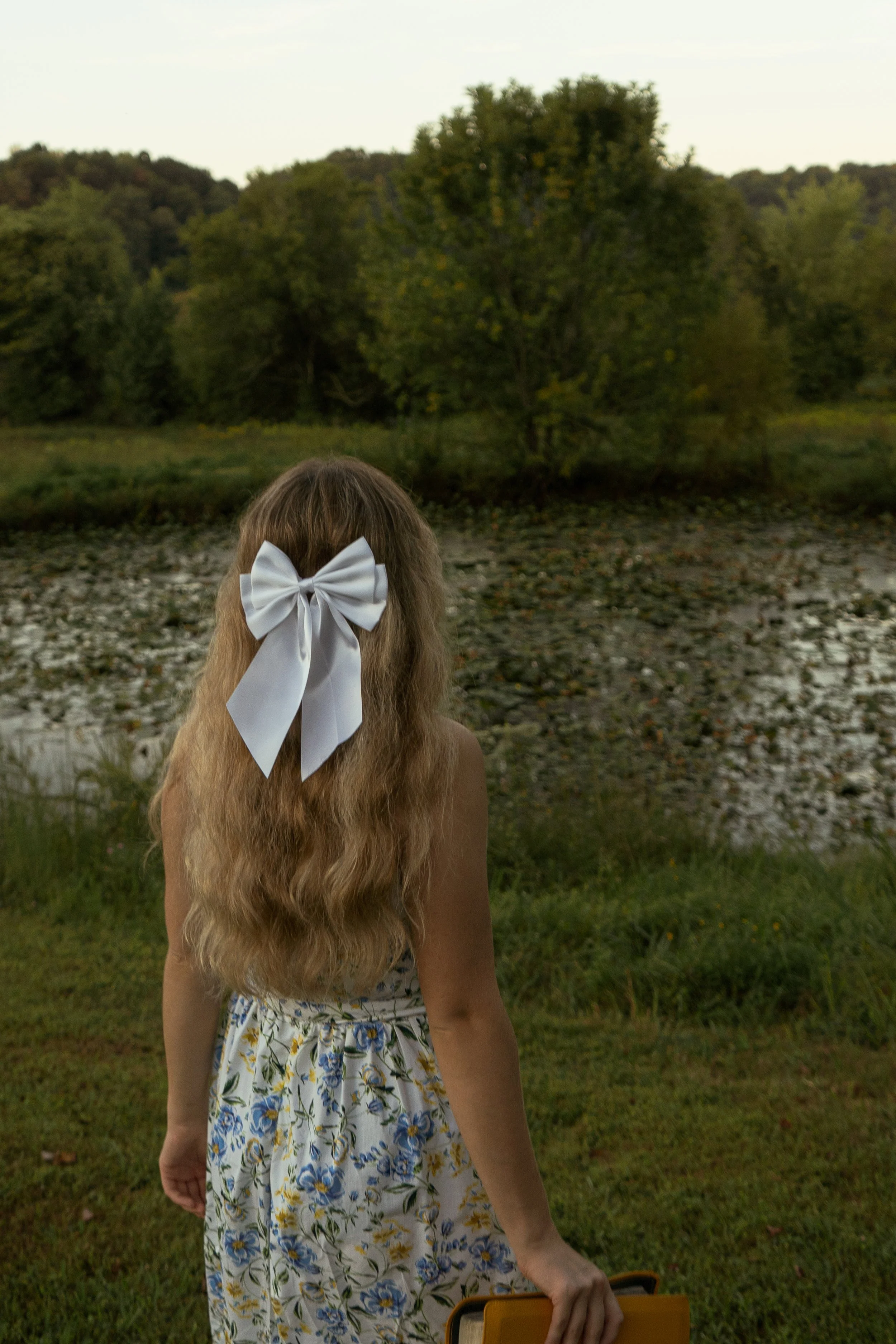 A girl with long curly hair tied with a white bow, wearing a floral dress, standing outdoors near a grassy area with a rocky riverbank and trees in the background.