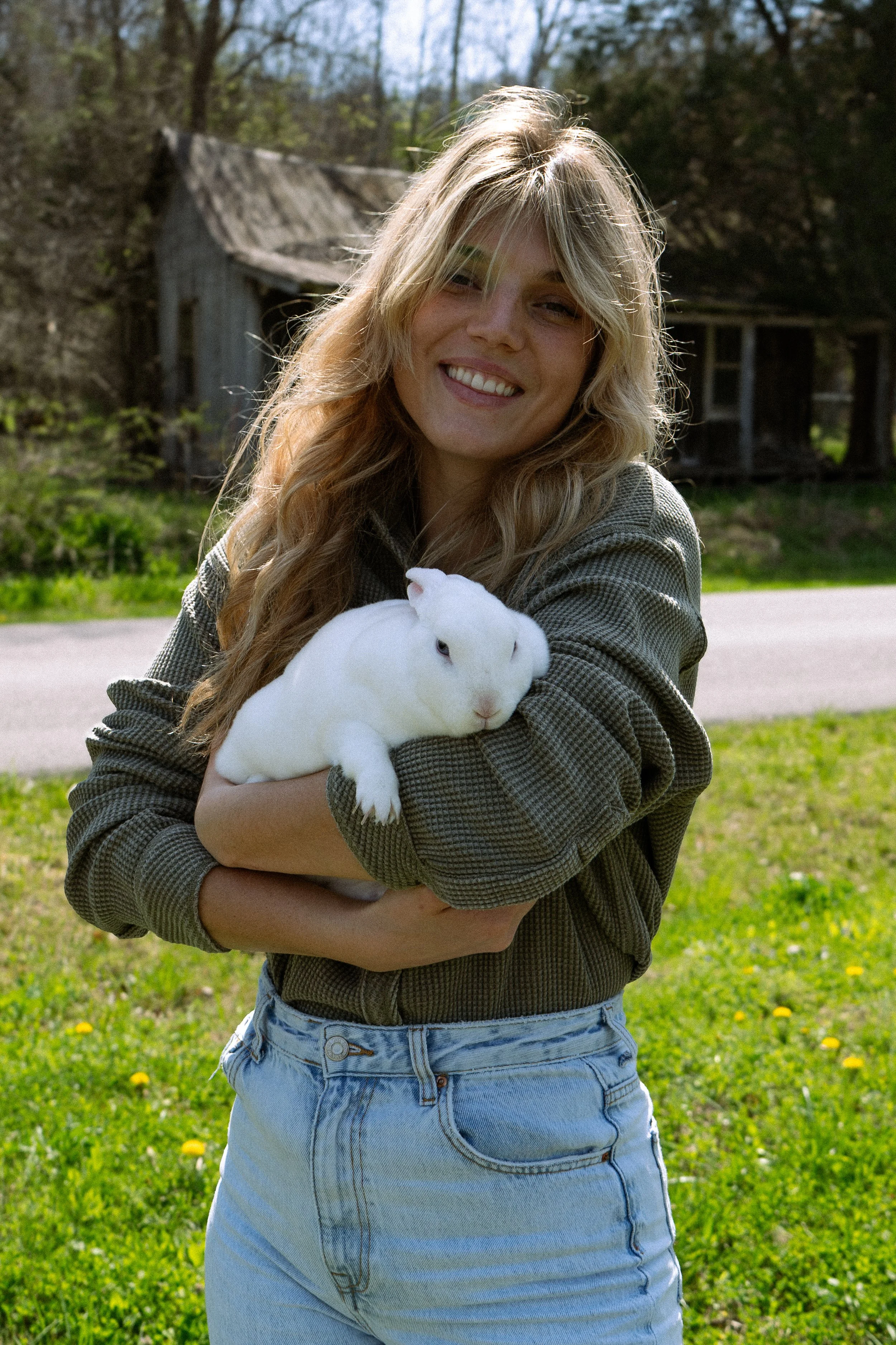 A young woman smiling and holding a white rabbit outdoors in a grassy area with trees and a rustic house in the background.
