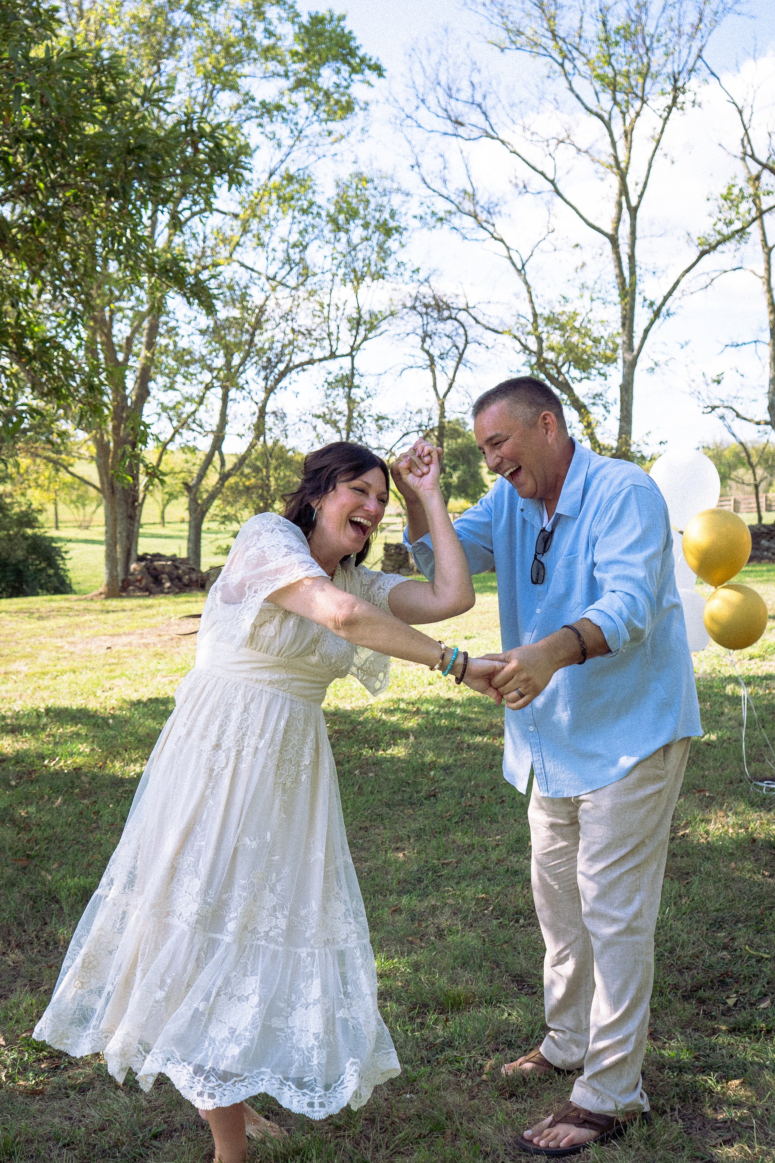A woman in a white lace dress and a man in a blue shirt and beige pants are outdoors, smiling and holding hands, with balloons in the background.