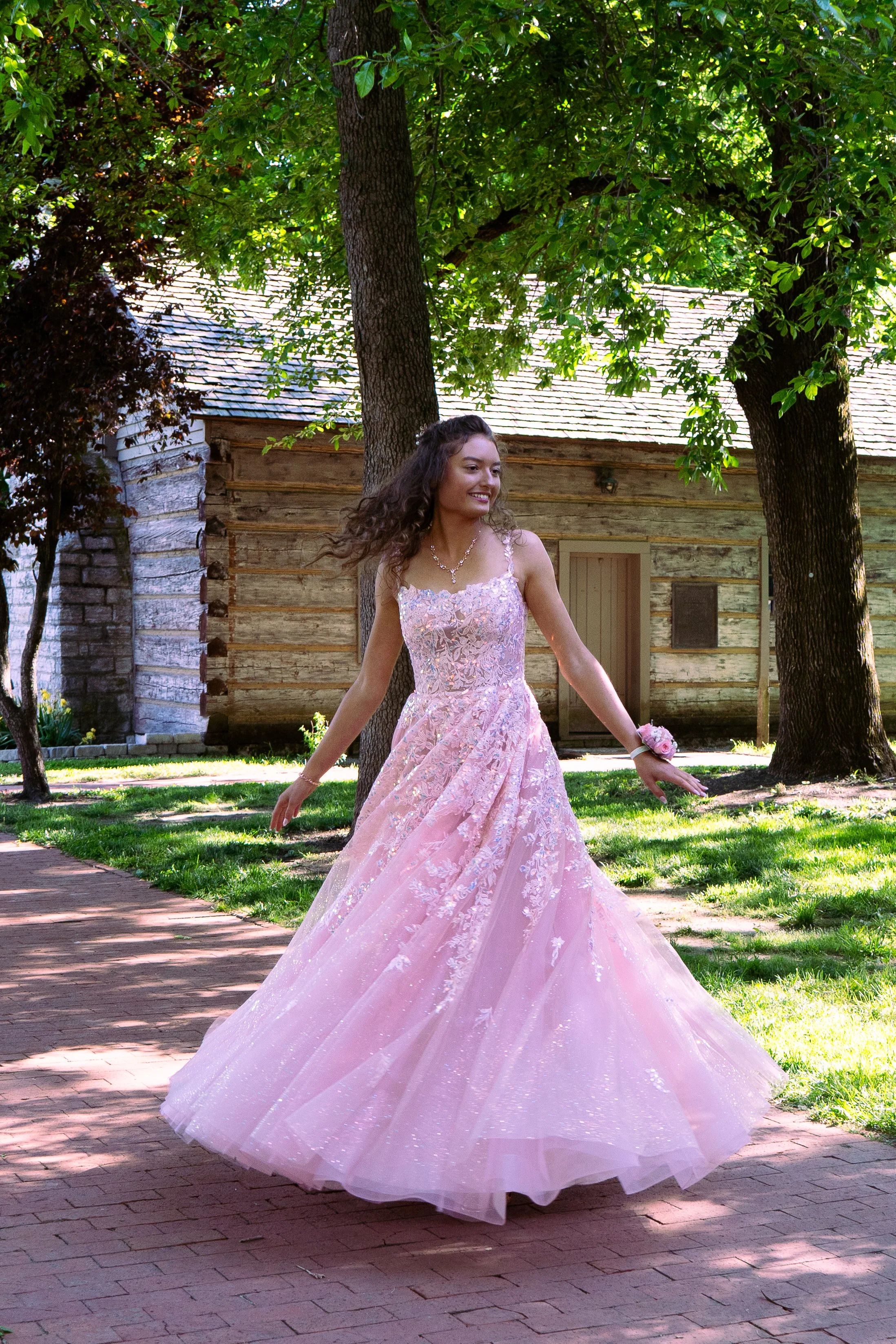A woman in a pink, lace wedding dress twirling outdoors near a large tree with green leaves, wooden house in the background, on a brick pathway surrounded by green grass.