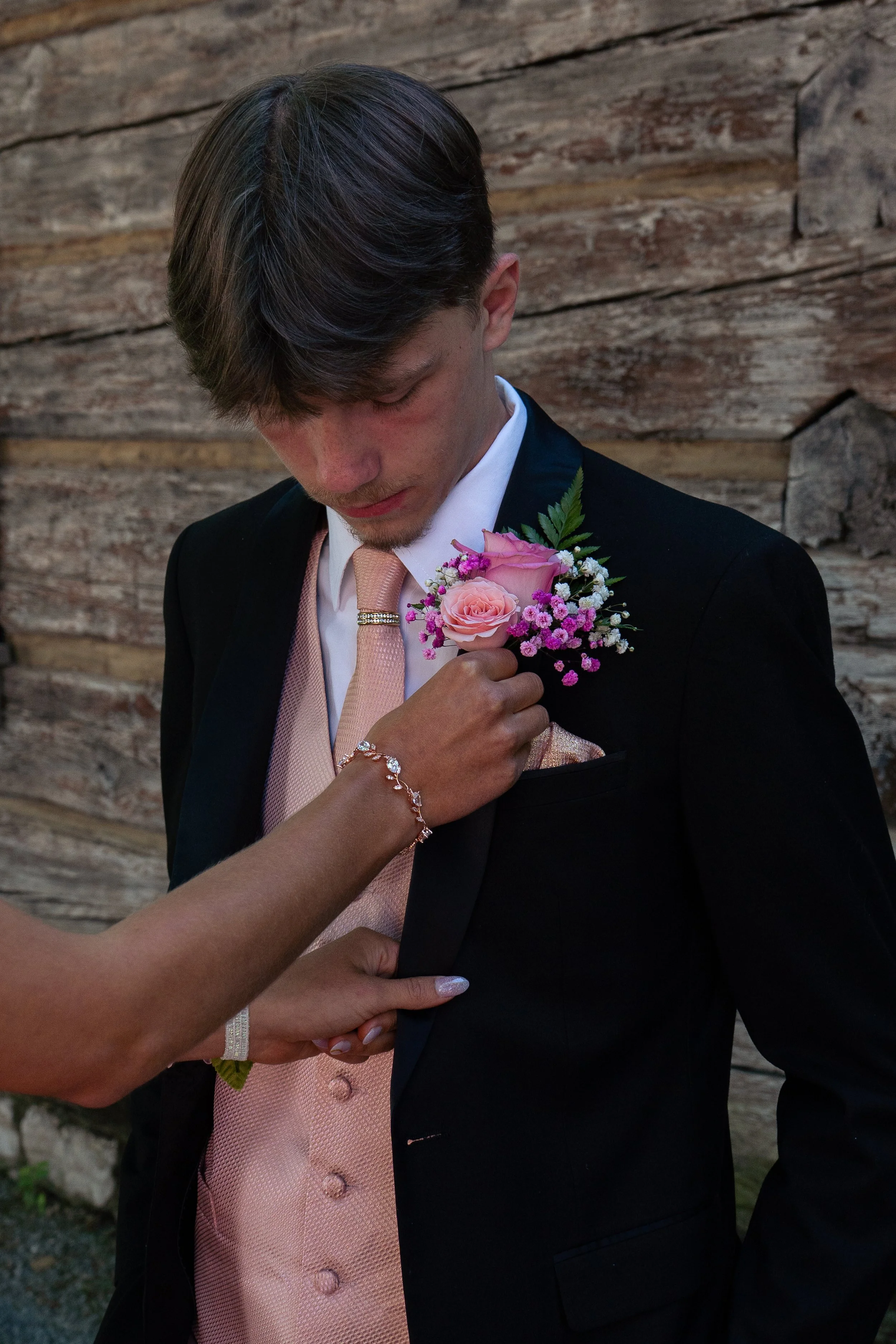 A young man in formal attire with a pale pink tie and a flower boutonniere on his suit lapel, standing outdoors against a wooden background, while a person adjusts his pink satin pocket square.
