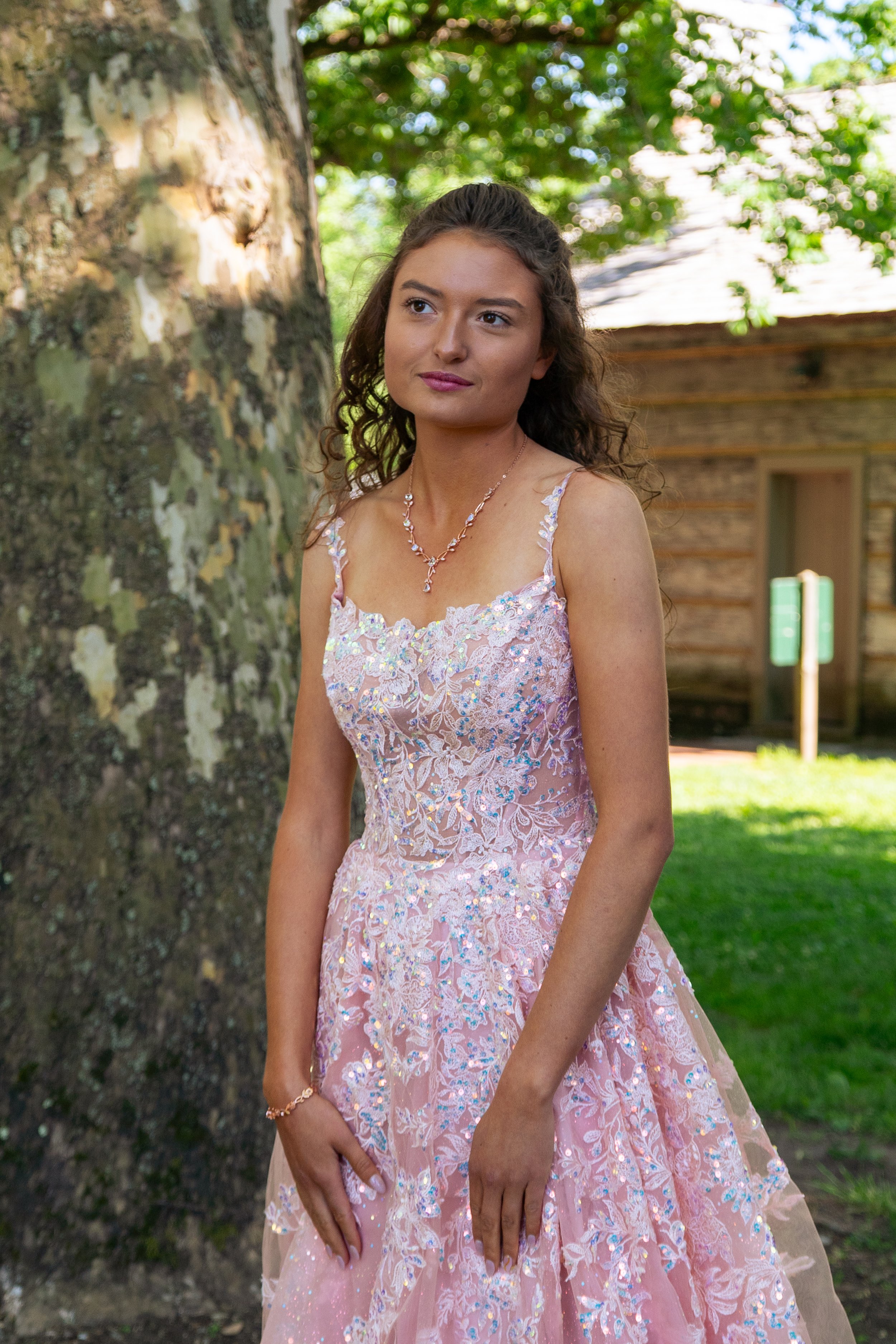 A young woman in a pink, sparkly, lacy evening gown standing outdoors near a tree in a park-like setting with a wooden building in the background.