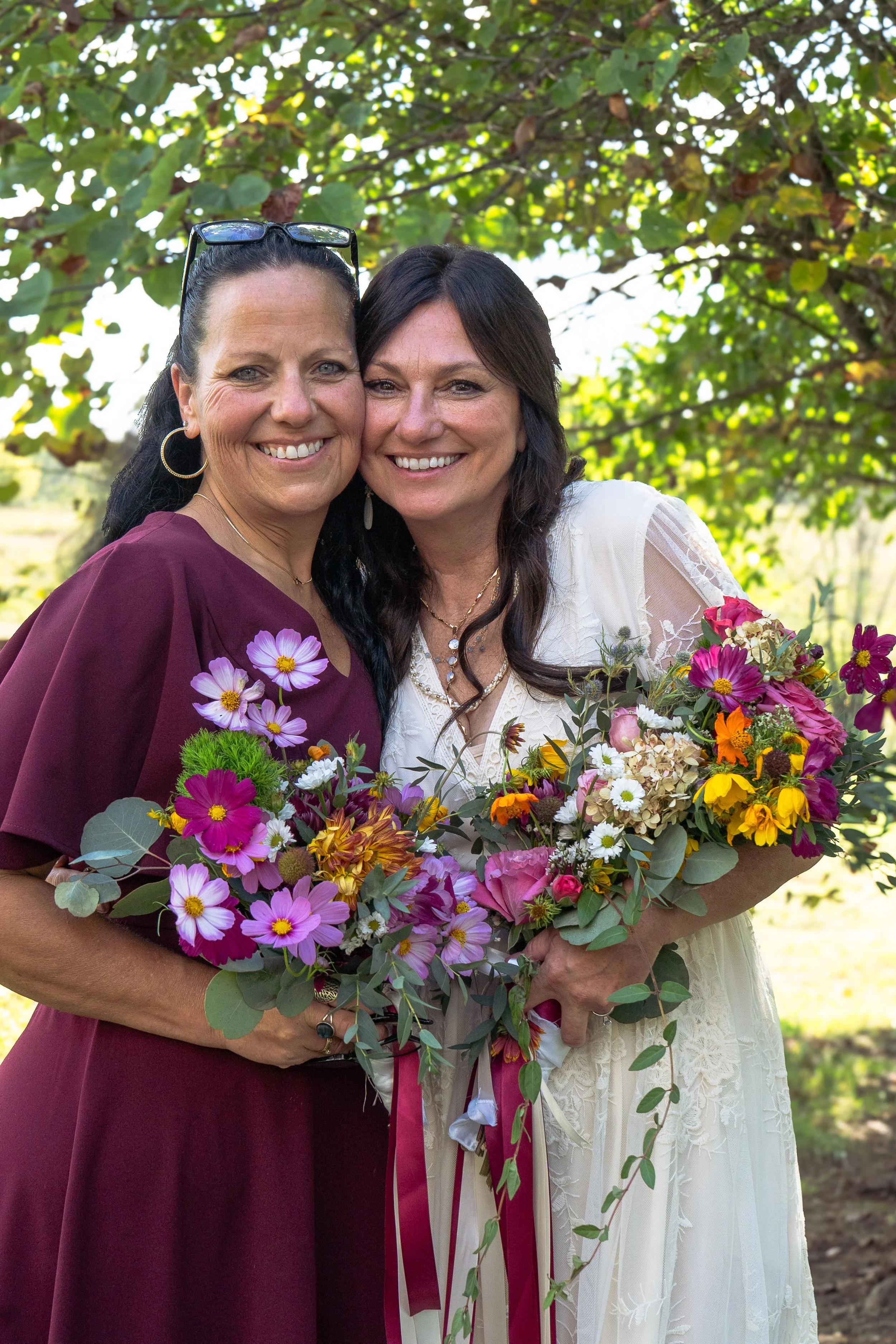 Two women smiling and embracing, holding colorful bouquets of flowers in front of a tree with green leaves.