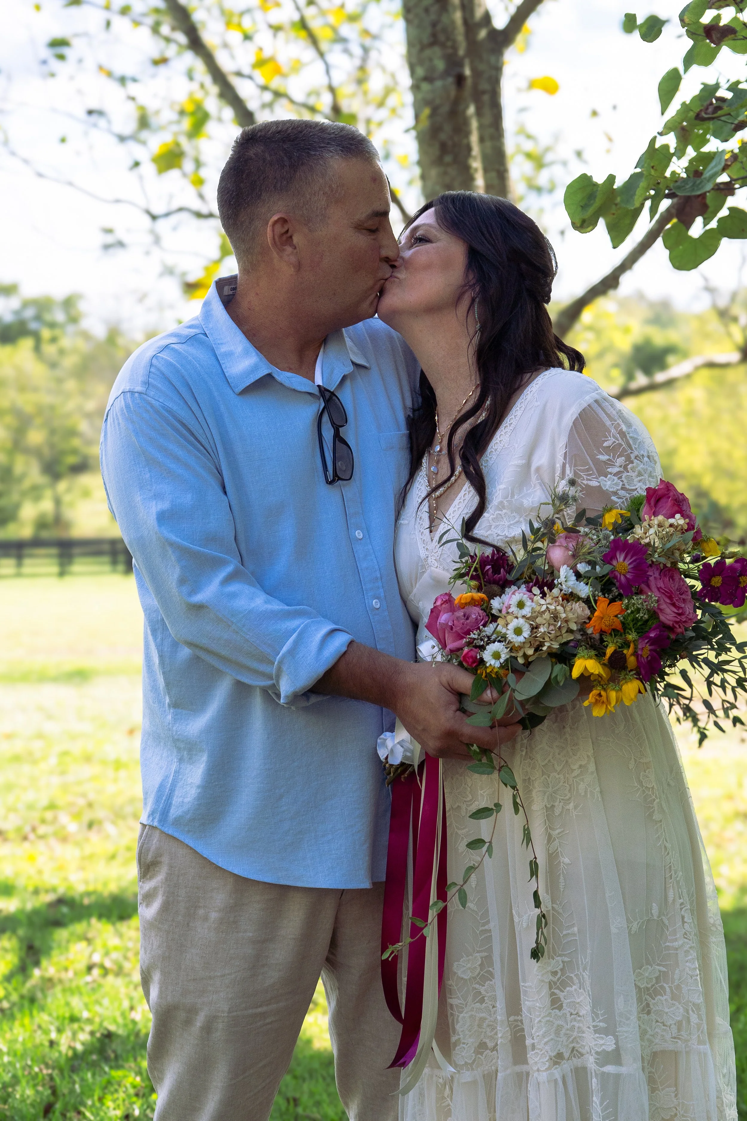 A couple dressed in wedding attire sharing a kiss outdoors on a sunny day, holding a large bouquet of colorful flowers with green foliage, with trees and bright sky in the background.