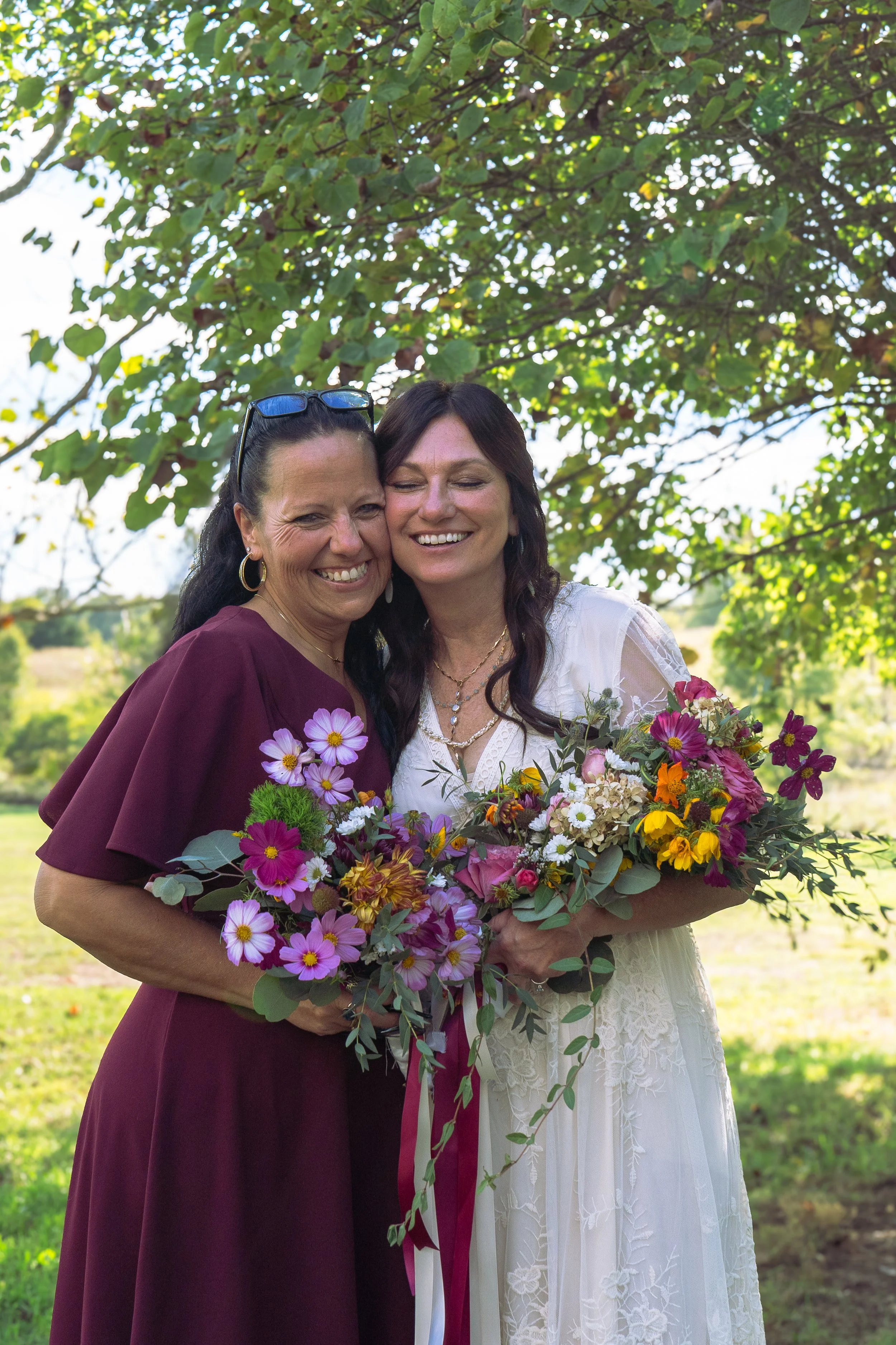 Two women smiling, hugging, and holding a large bouquet of colorful flowers outdoors under green leafy trees. One woman has dark hair and wears a dark purple dress with sunglasses on her head; the other has brunette hair and wears a white lace dress.
