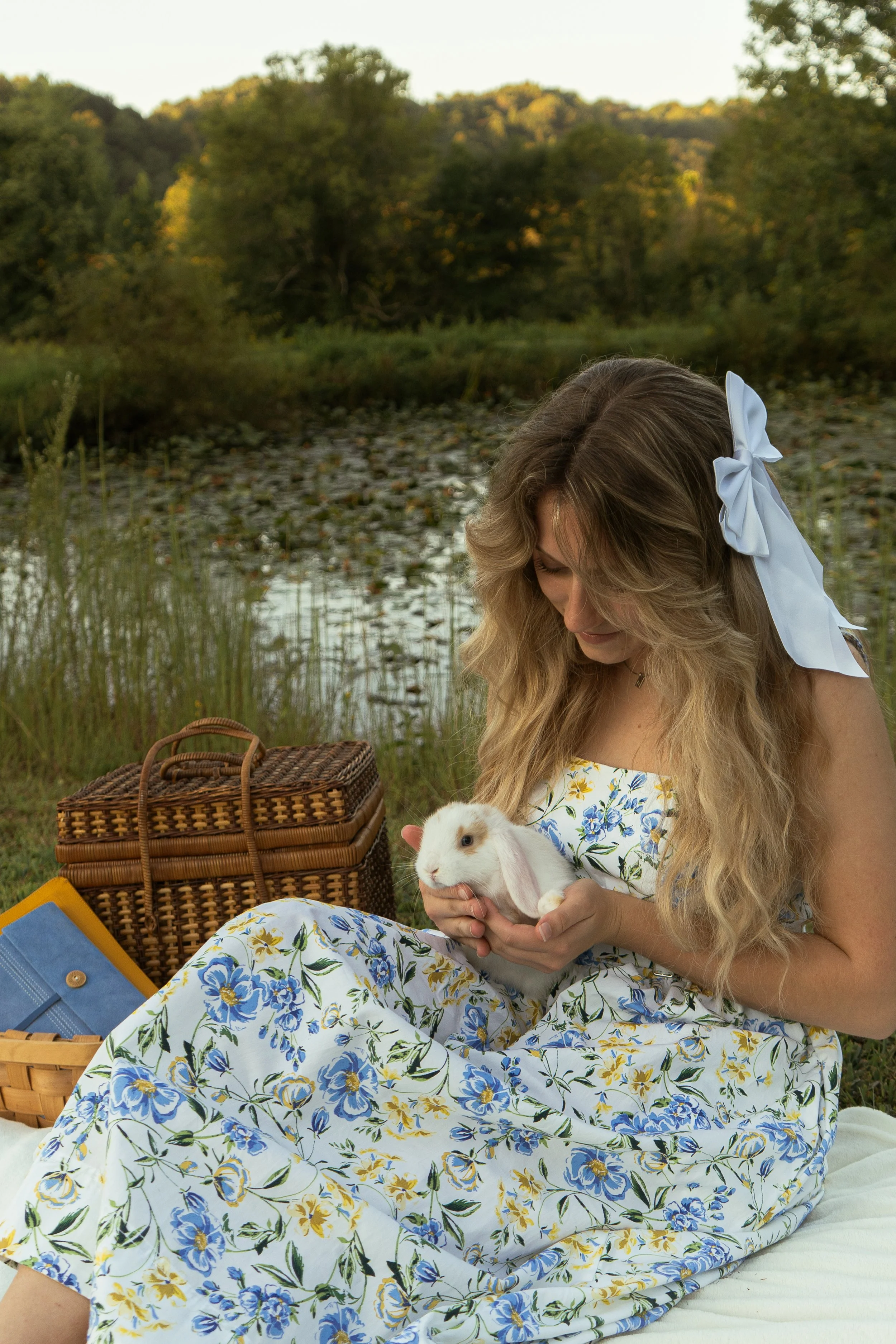 A woman with long, wavy hair and a large white bow in her hair is sitting outdoors near a body of water surrounded by trees. She is holding a white and brown rabbit and smiling while looking down. She is wearing a white dress with a blue and yellow f