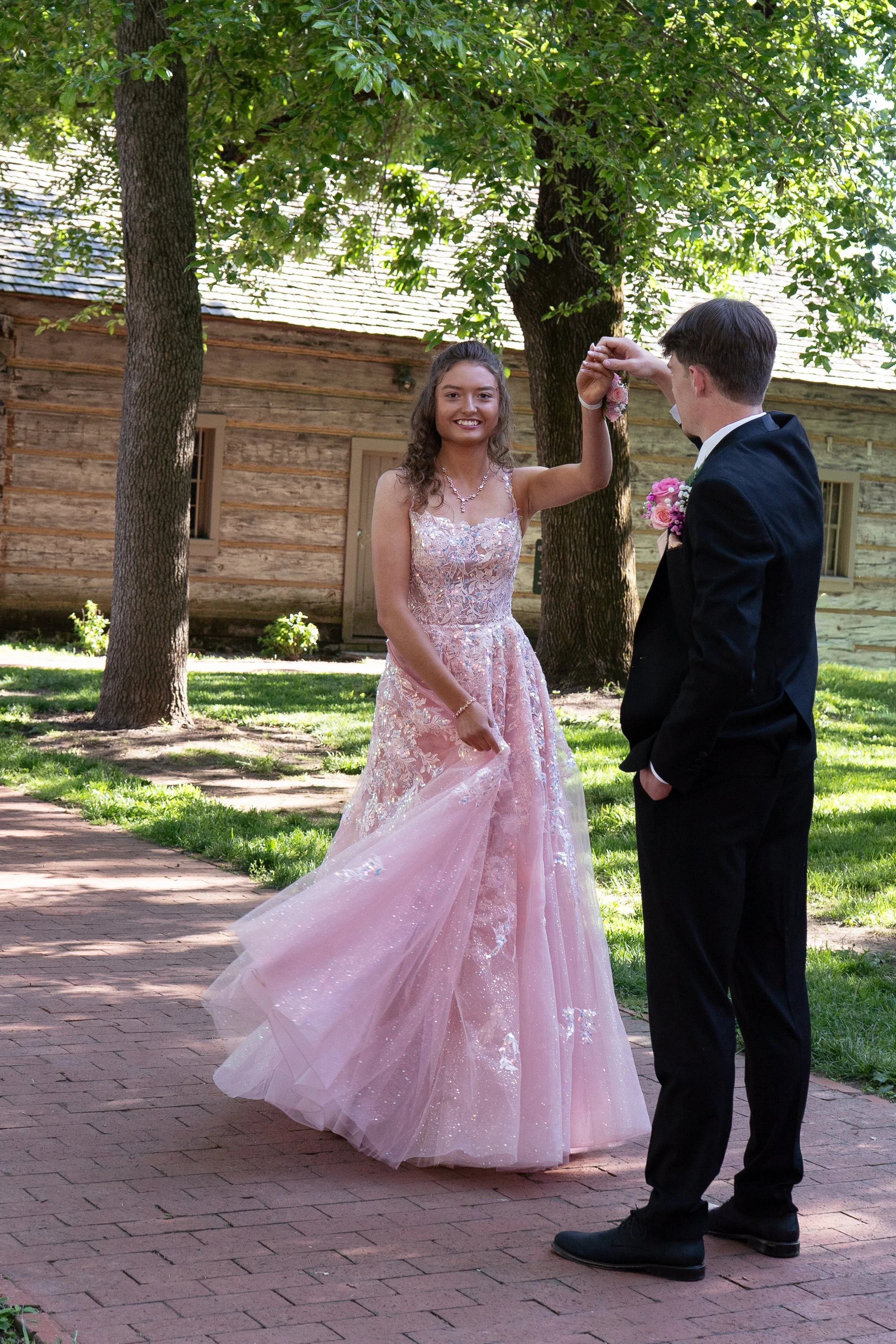 A couple dancing outdoors on a brick pathway, with the woman in a pink, embellished gown and the man in a dark suit, surrounded by trees and a wooden building.