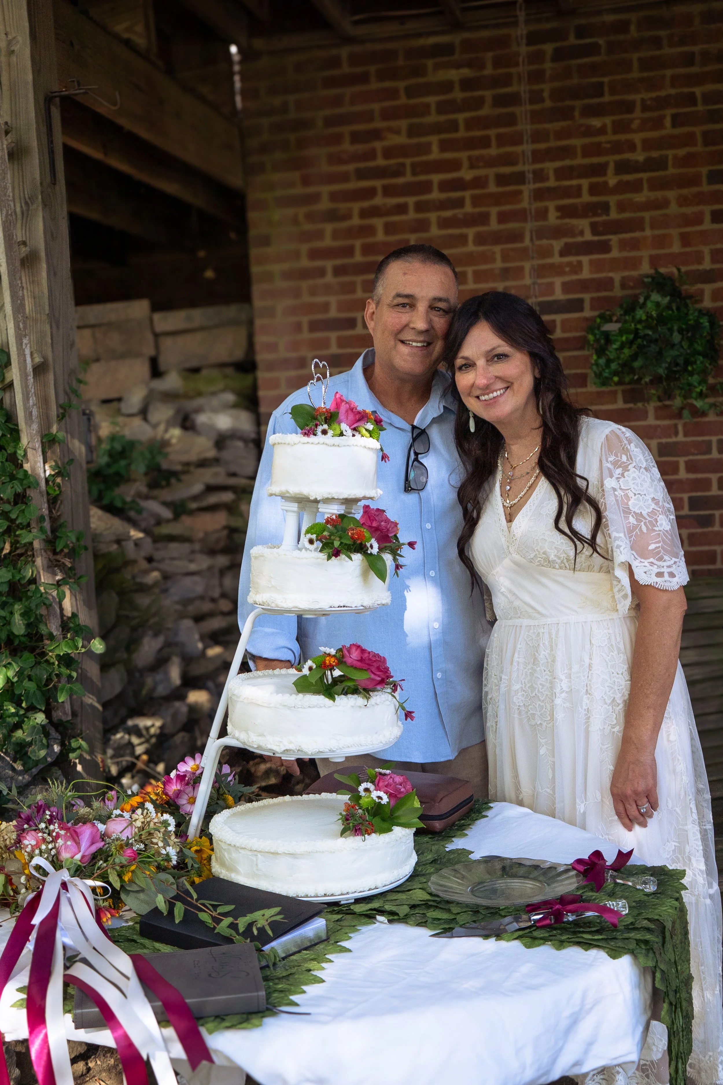 A smiling couple, dressed in wedding attire, stands outdoors next to a tiered wedding cake decorated with flowers. The table in front of them is covered with greenery, flowers, and ribbons, with a brick wall in the background.