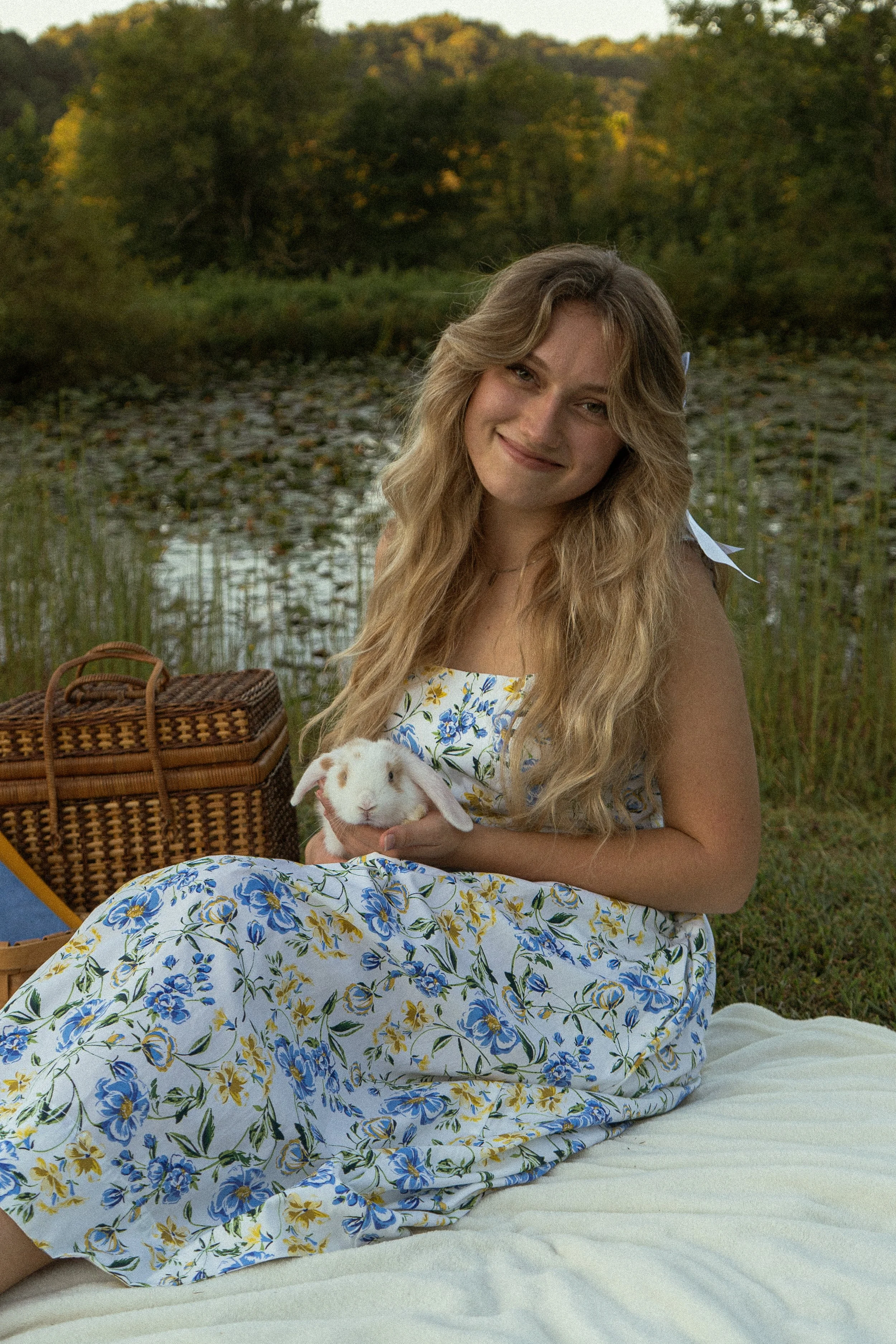 A young woman with long wavy blonde hair sitting outdoors by a lake, holding a white rabbit with brown spots, smiling at the camera. She's wearing a white dress with blue and yellow floral pattern, and there's a wicker picnic basket beside her. The b