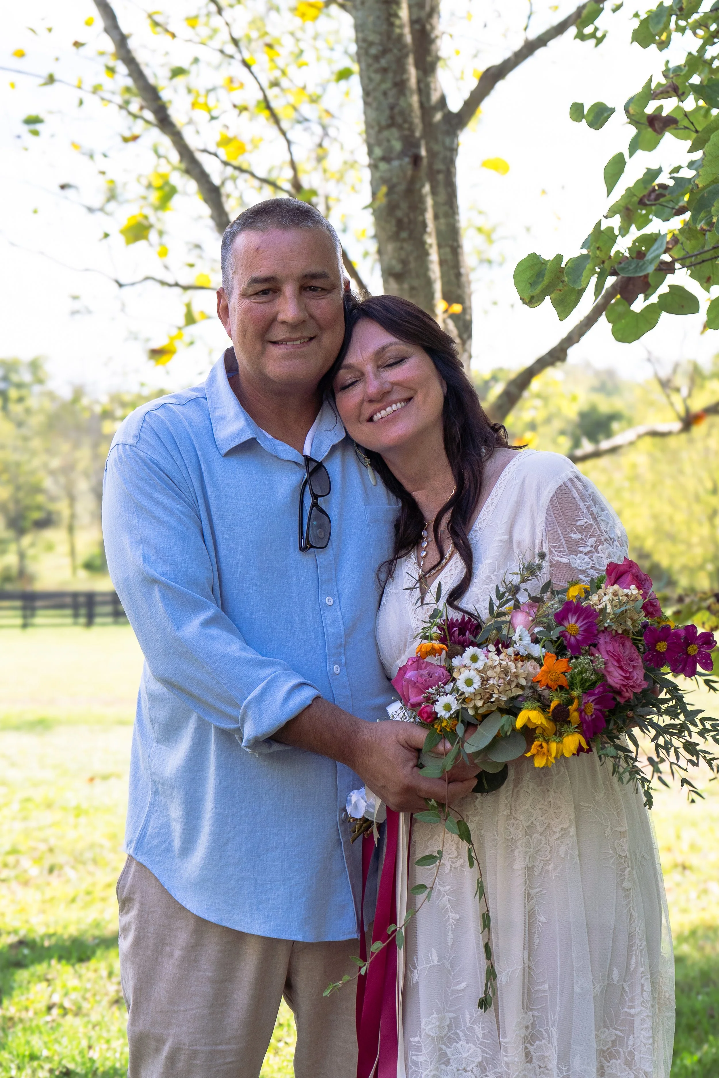 A smiling couple, a man and a woman, standing outdoors in a park-like setting with green grass, trees, and a wooden fence in the background. The woman is holding a large bouquet of colorful flowers. They are embracing each other with their heads clos