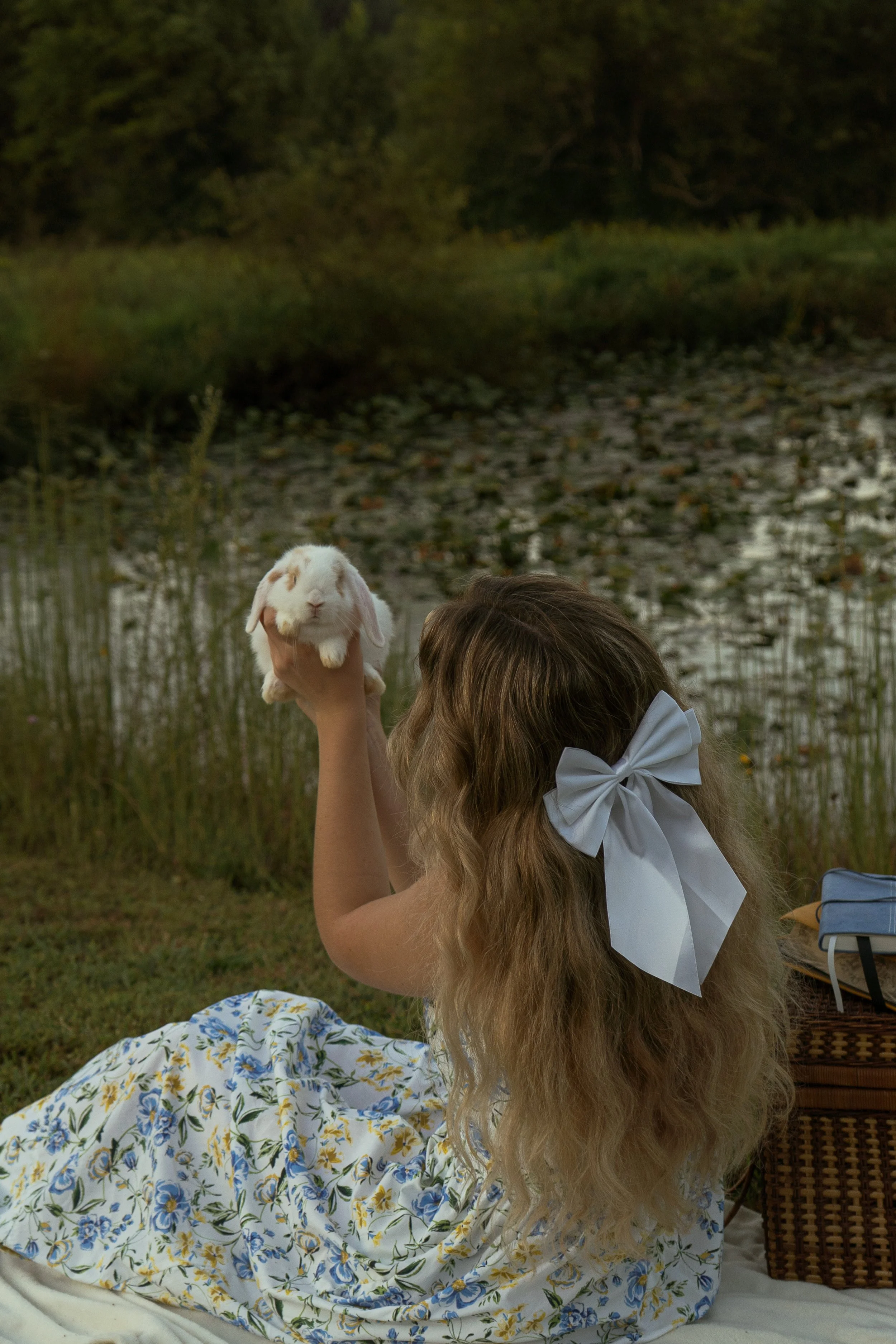 A person with long curly blonde hair, wearing a large white bow, is holding a white rabbit near a lake with trees and lily pads in the background during sunset.