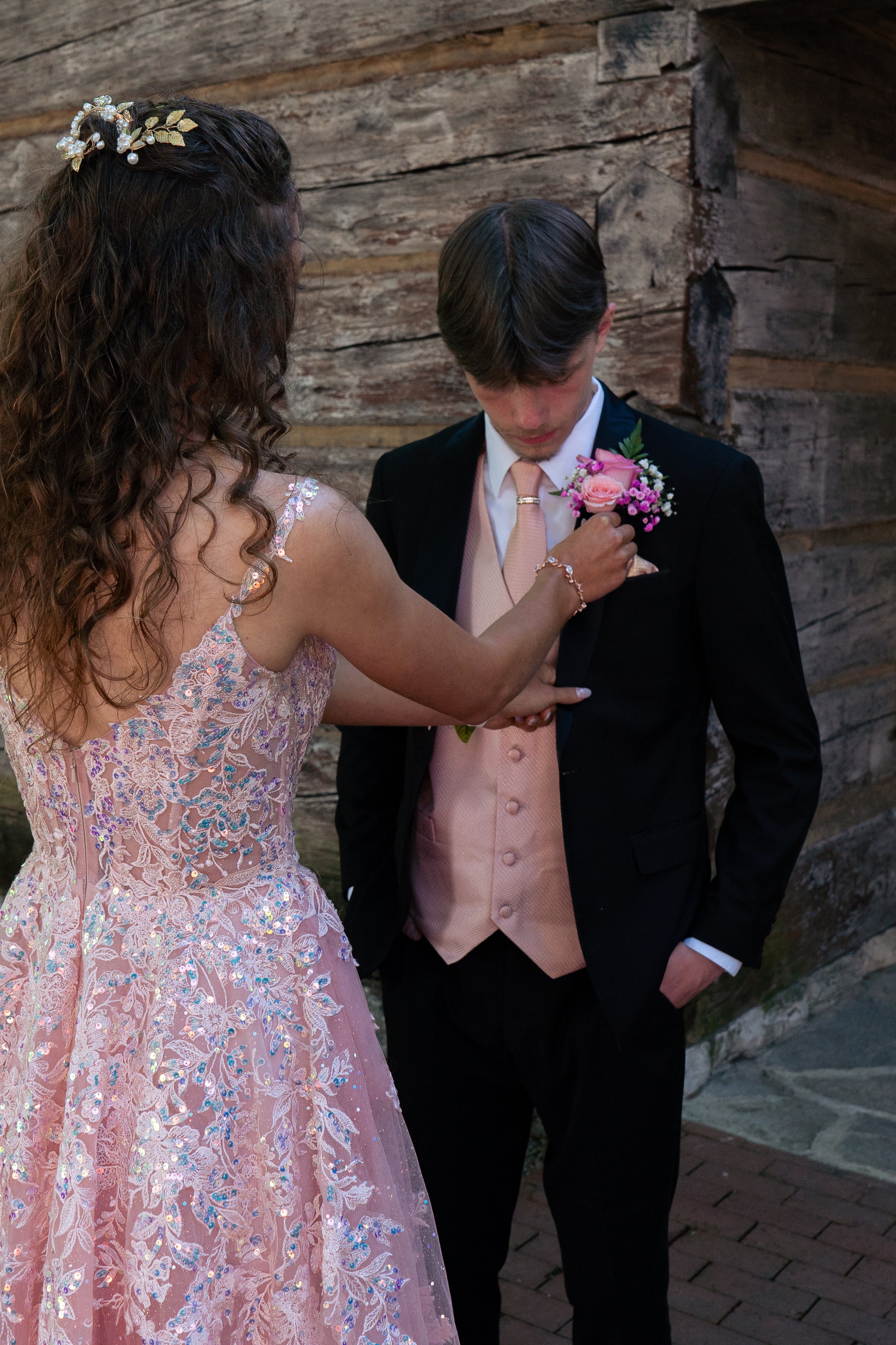 A woman in a pink lace dress with sequins helps a young man in a black suit with a pink tie and matching vest put on a boutonniere.