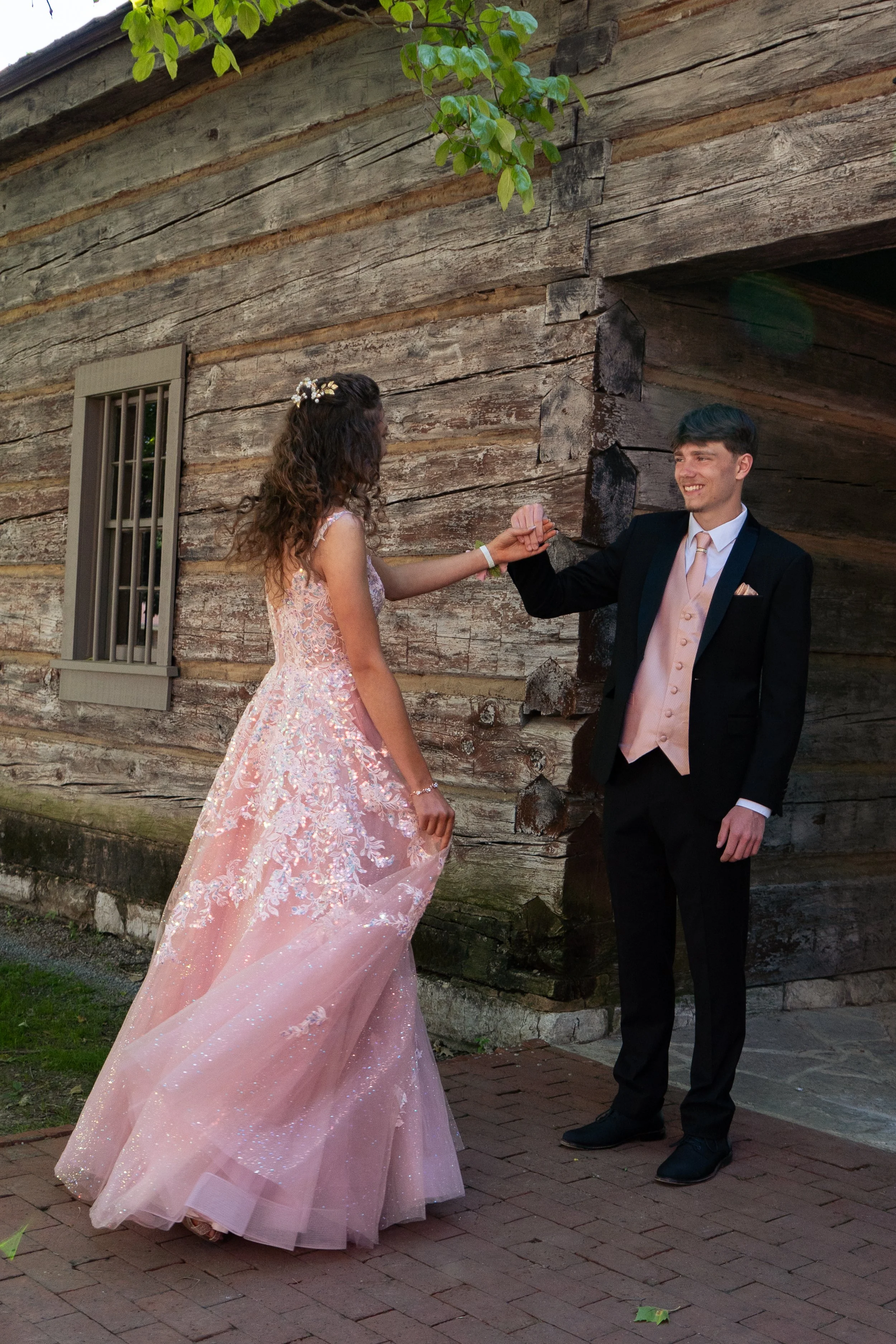 A young woman in a light pink, sparkly ball gown with floral embroidery and a young man in a tuxedo with a peach vest and tie, standing outside in front of a rustic wooden building, holding hands and smiling.