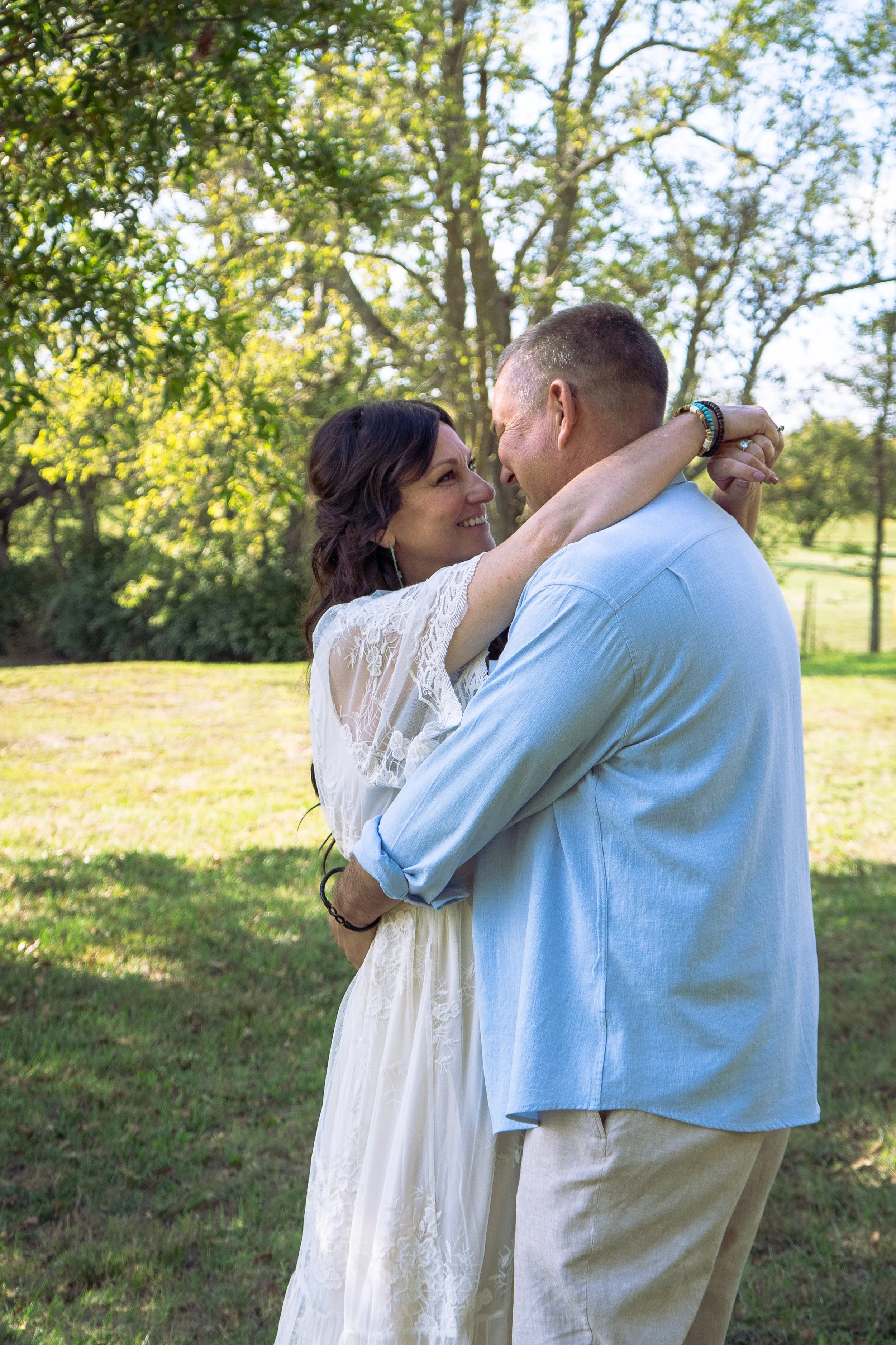 A couple dancing outdoors on a sunny day, surrounded by green trees, with the woman wearing a white lace dress and the man in a light blue shirt.
