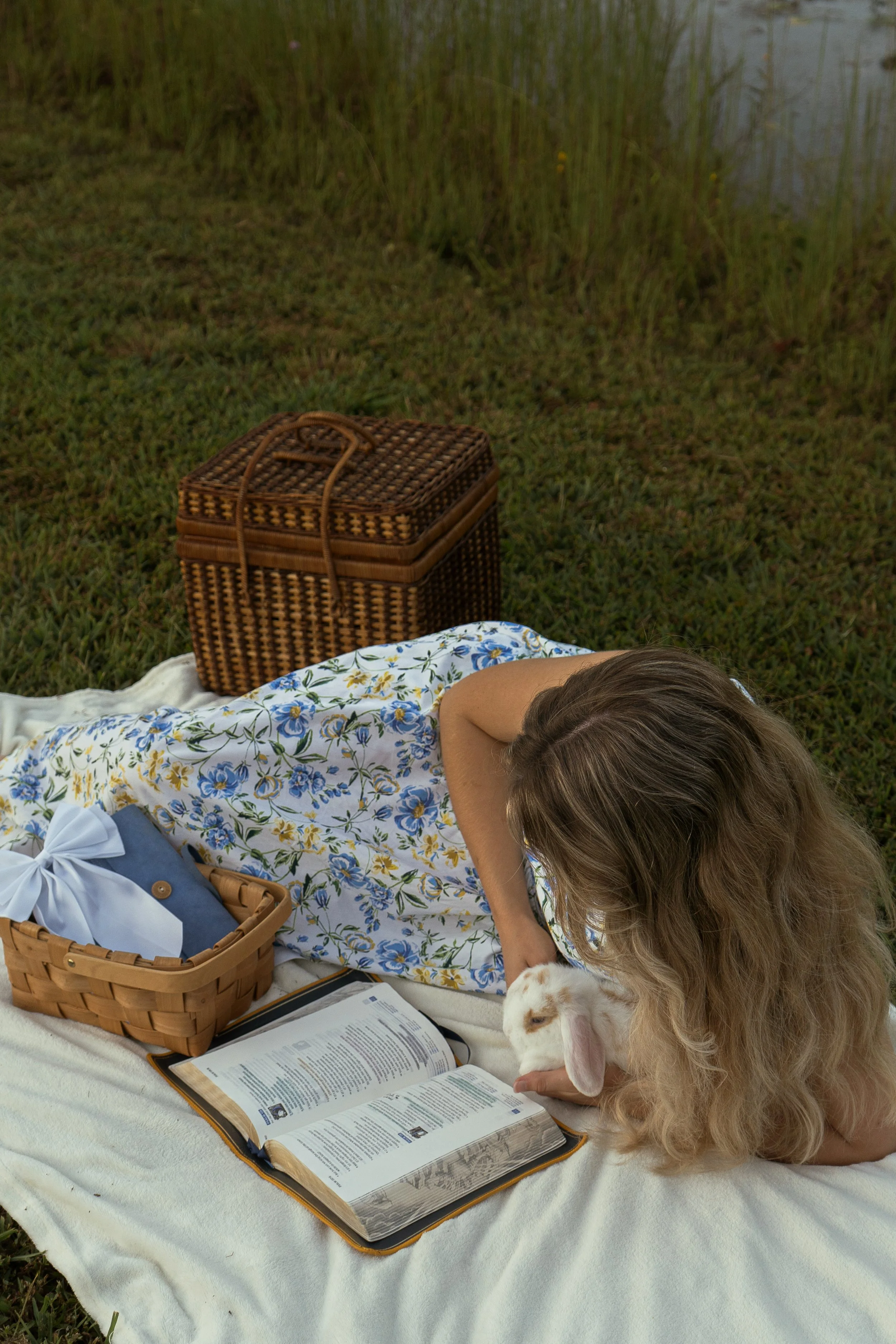 A person lying on a blanket outdoors near a body of water, reading a book, with a rabbit nearby. There is a woven picnic basket and a wicker box on the blanket.