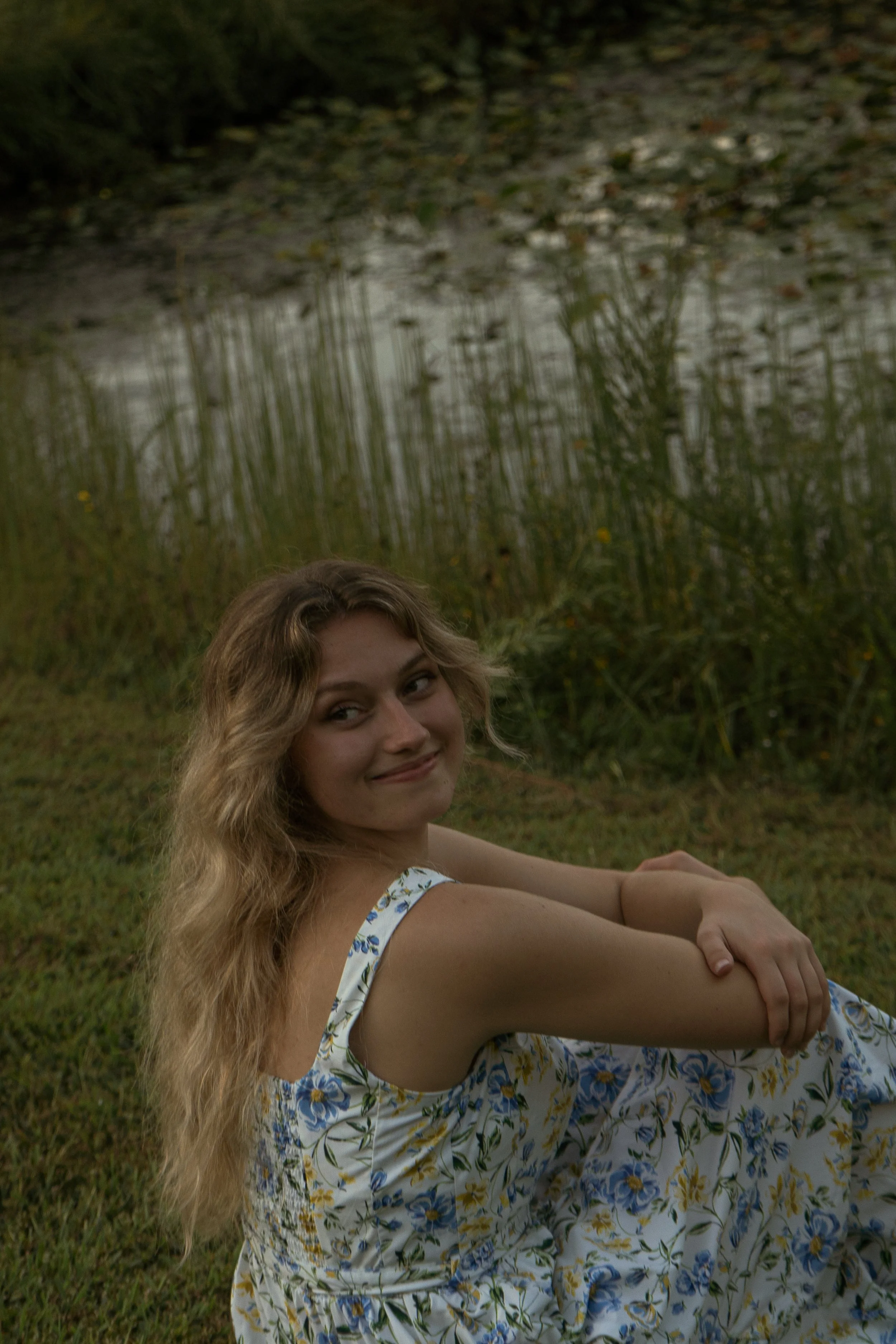 A young woman with long, wavy blonde hair sitting outside on grass, smiling at the camera, wearing a sleeveless dress with a floral print, in a natural setting with greenery and a wet, muddy area in the background.