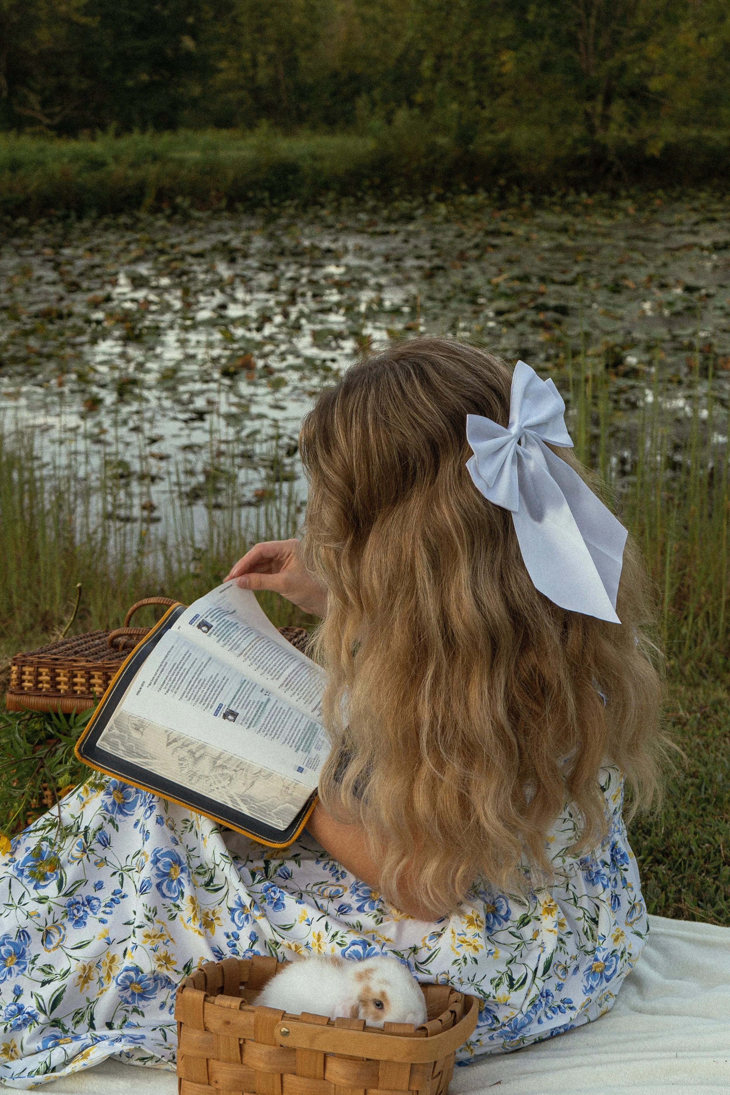 A woman with long, wavy blonde hair, wearing a large white bow, reading a magazine outdoors near a pond with lily pads. She is dressed in a floral dress with blue and yellow flowers. A small, fluffy rabbit with brown and white fur is sitting in a woo