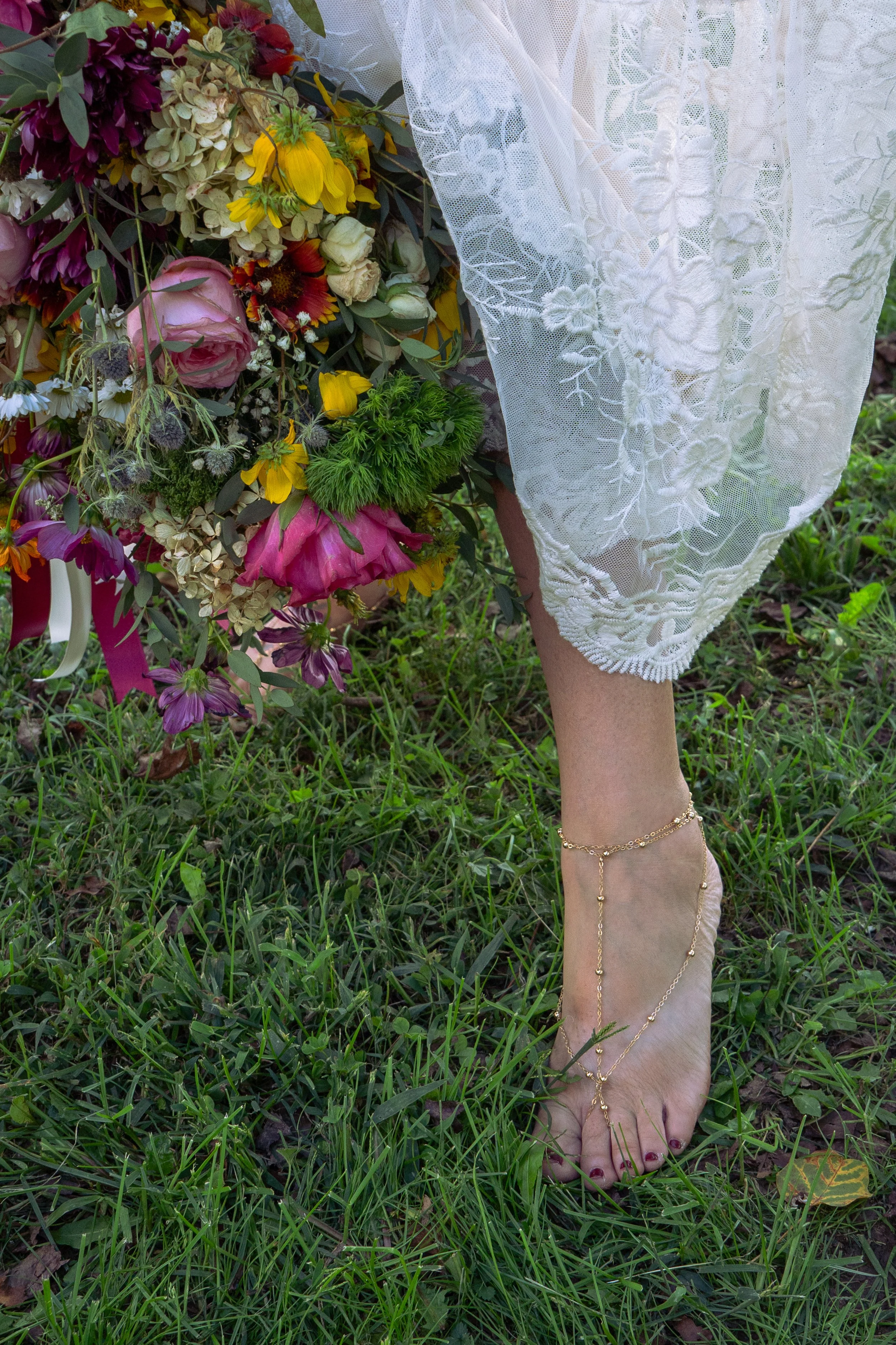 A person's bare foot with painted toenails wearing a gold ankle chain, standing on grass next to a bouquet of colorful flowers and a white lace fabric.