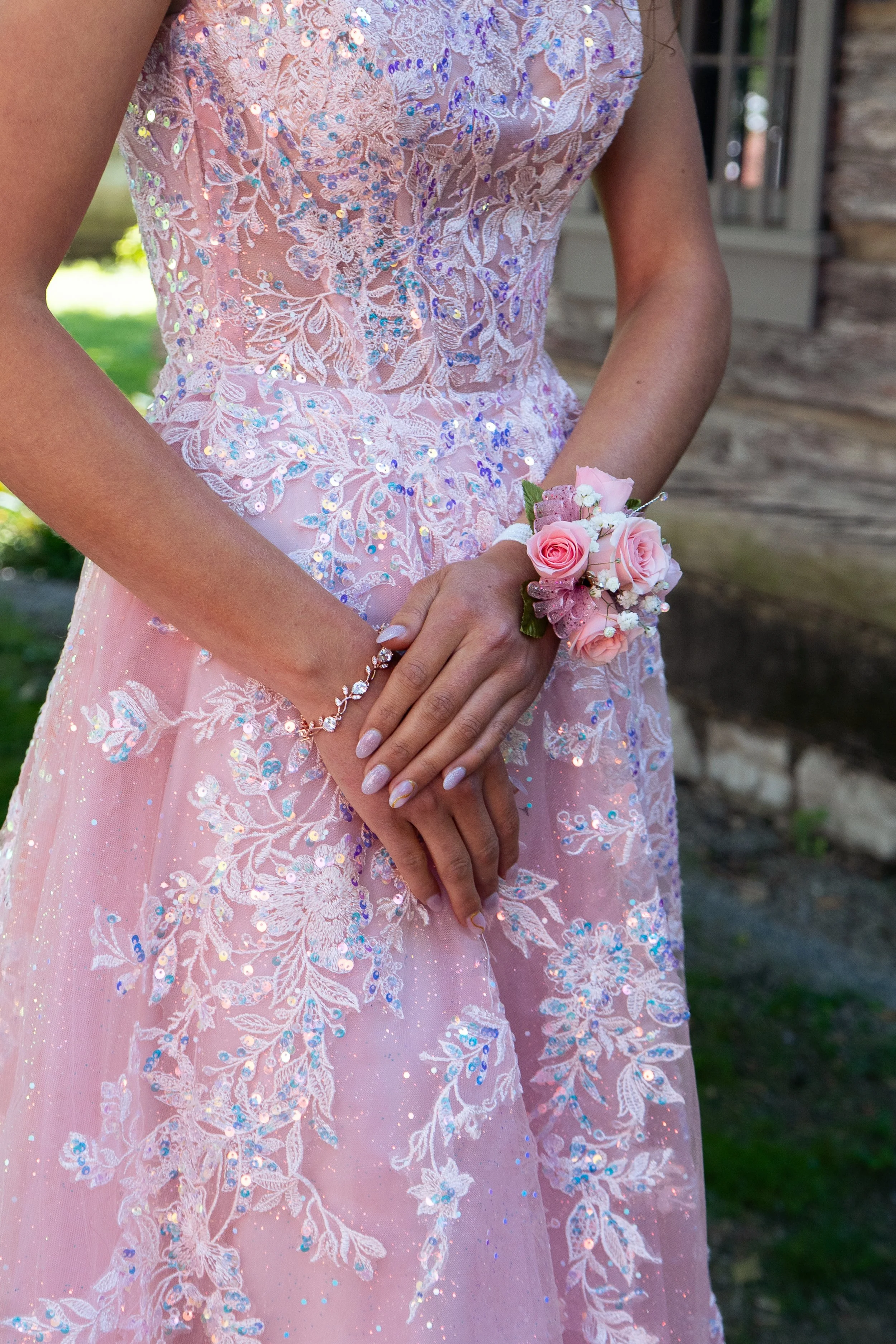 A woman in a pink, sequined, embroidered dress holding a small bouquet of pink roses with white baby's breath. Her hands and part of her waist are visible against a rustic wooden building and green outdoor background.
