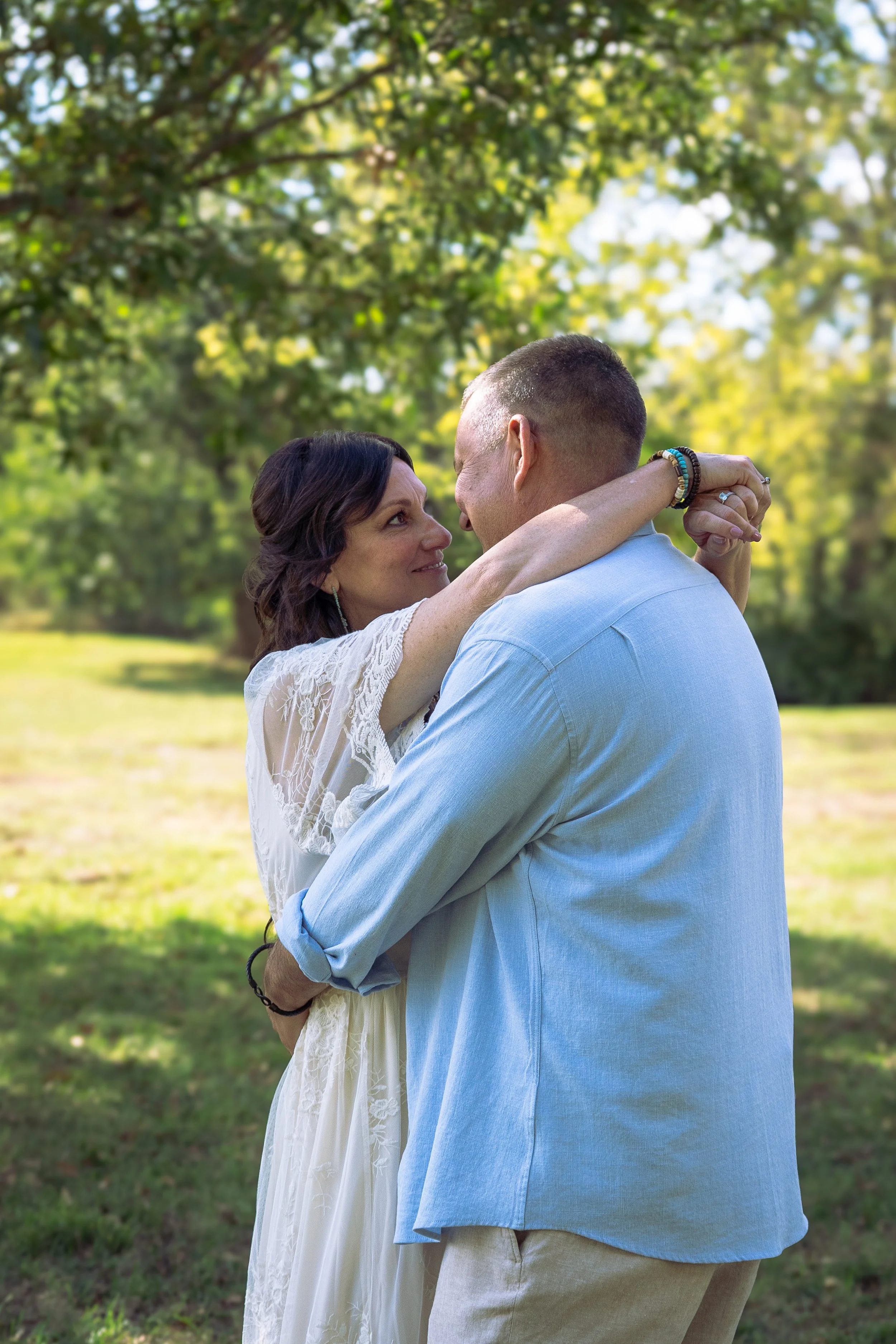 A couple is dancing outdoors in a lush green park, looking into each other's eyes with the woman smiling. The woman is wearing a white lace dress and has dark wavy hair, while the man is wearing a light blue shirt and has short hair. They are embraci