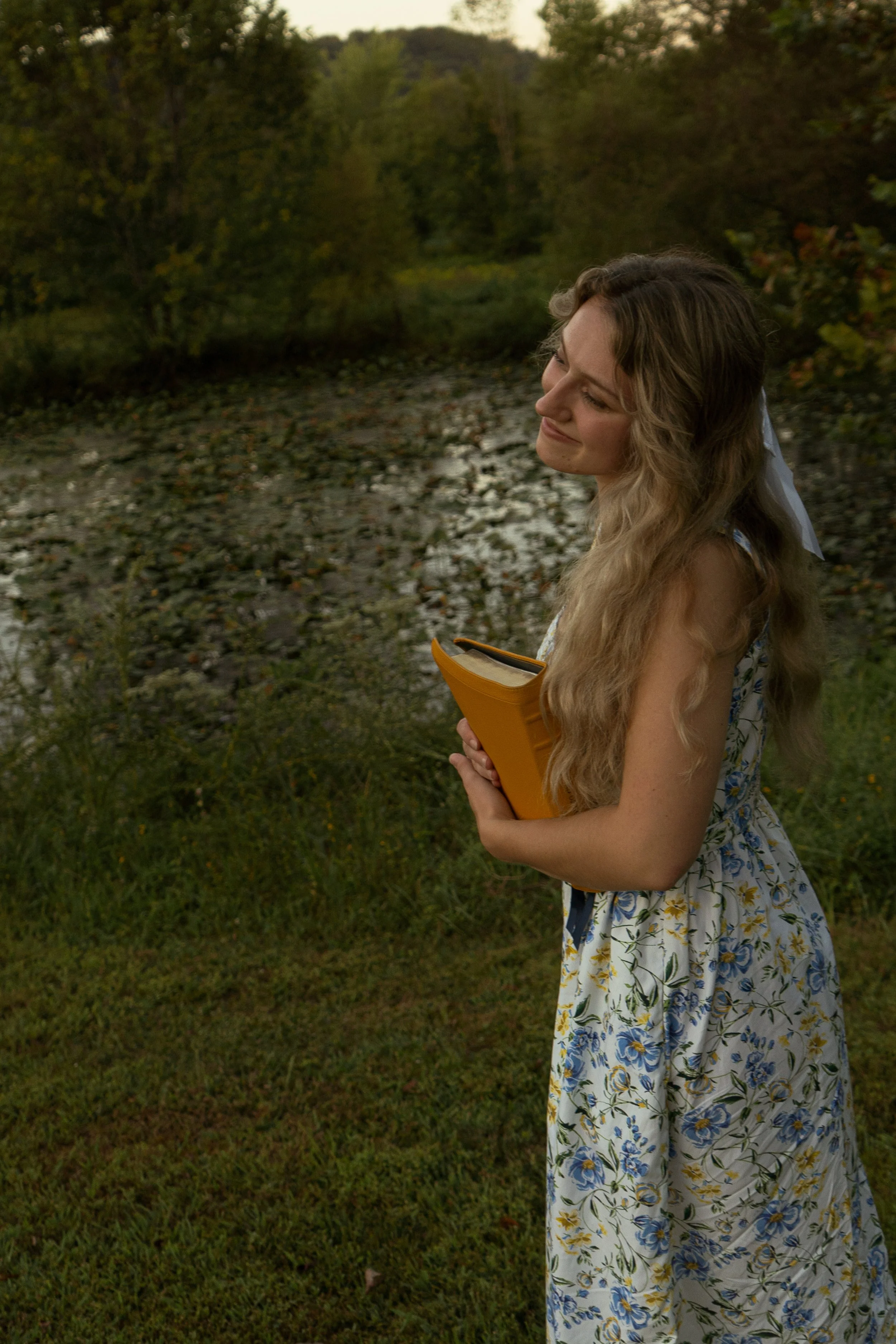 A young woman with long wavy hair holding a book, standing outdoors near a river with green trees, wearing a white floral dress, during the evening.