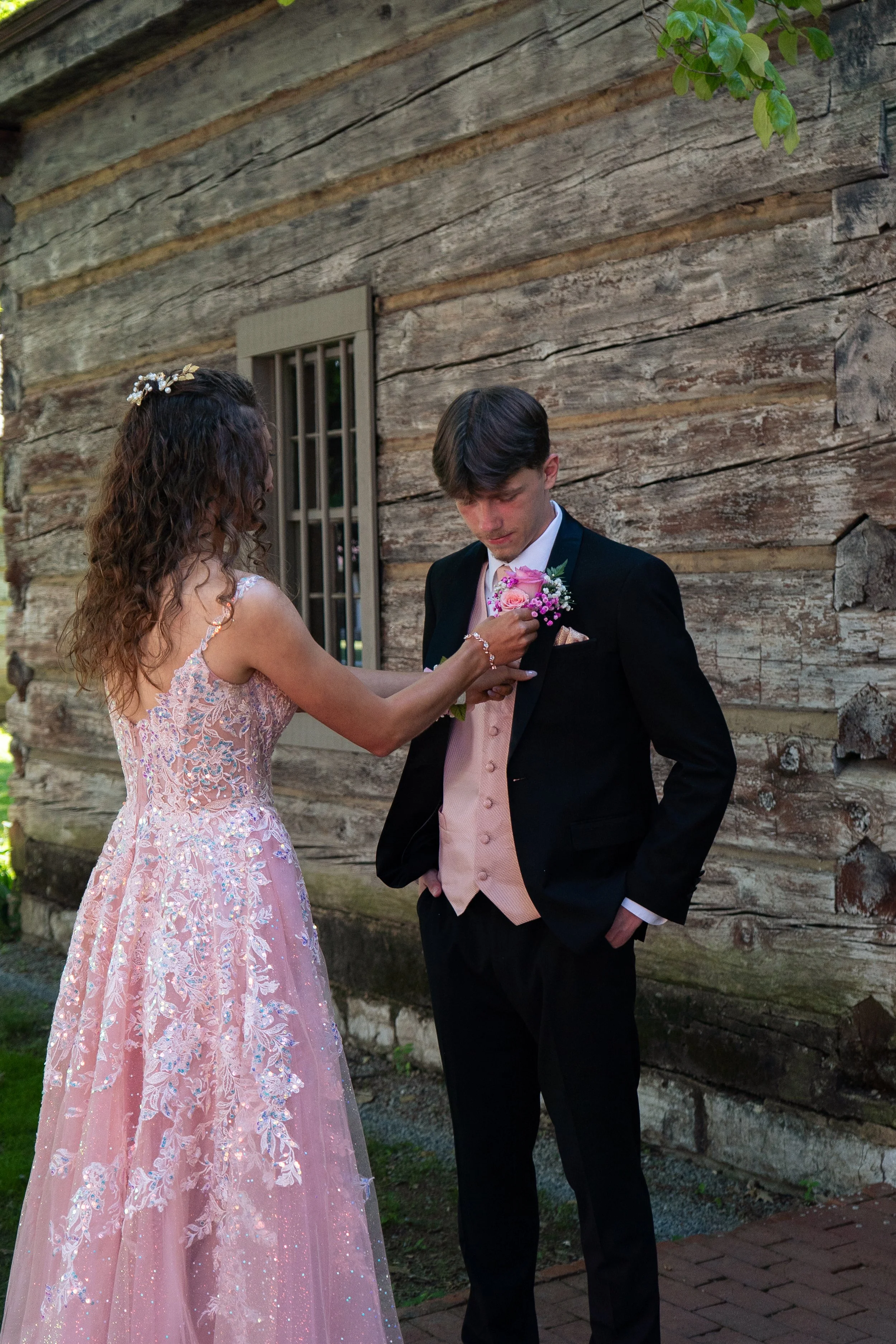 A woman in a pink lace dress pins a pink boutonniere onto a young man's black tuxedo, standing outside a rustic wooden building with a window and a brick pathway.