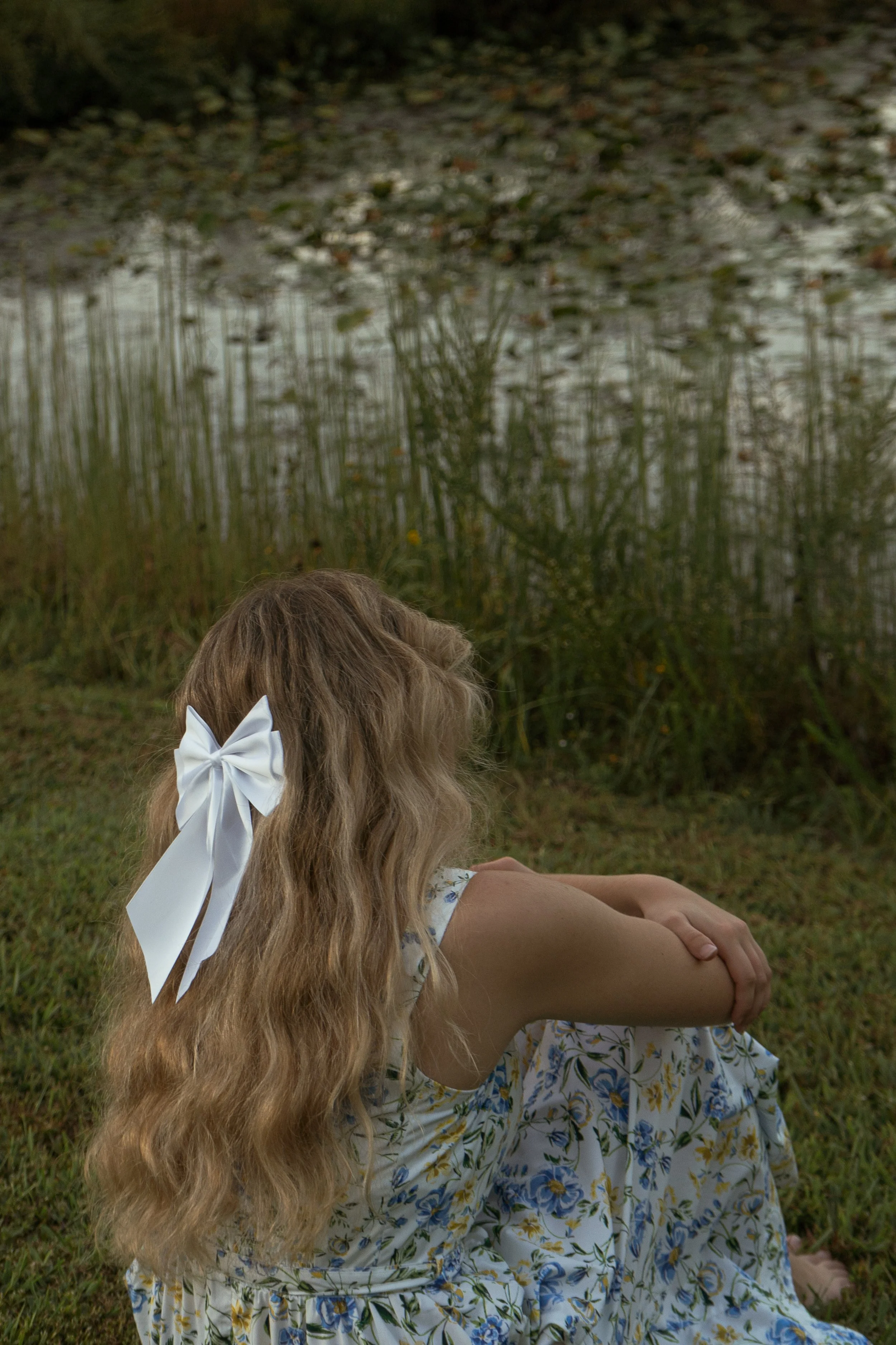 Girl with long blonde hair and a large white bow sitting on grass beside a water body with greenery and trees reflected in the water.