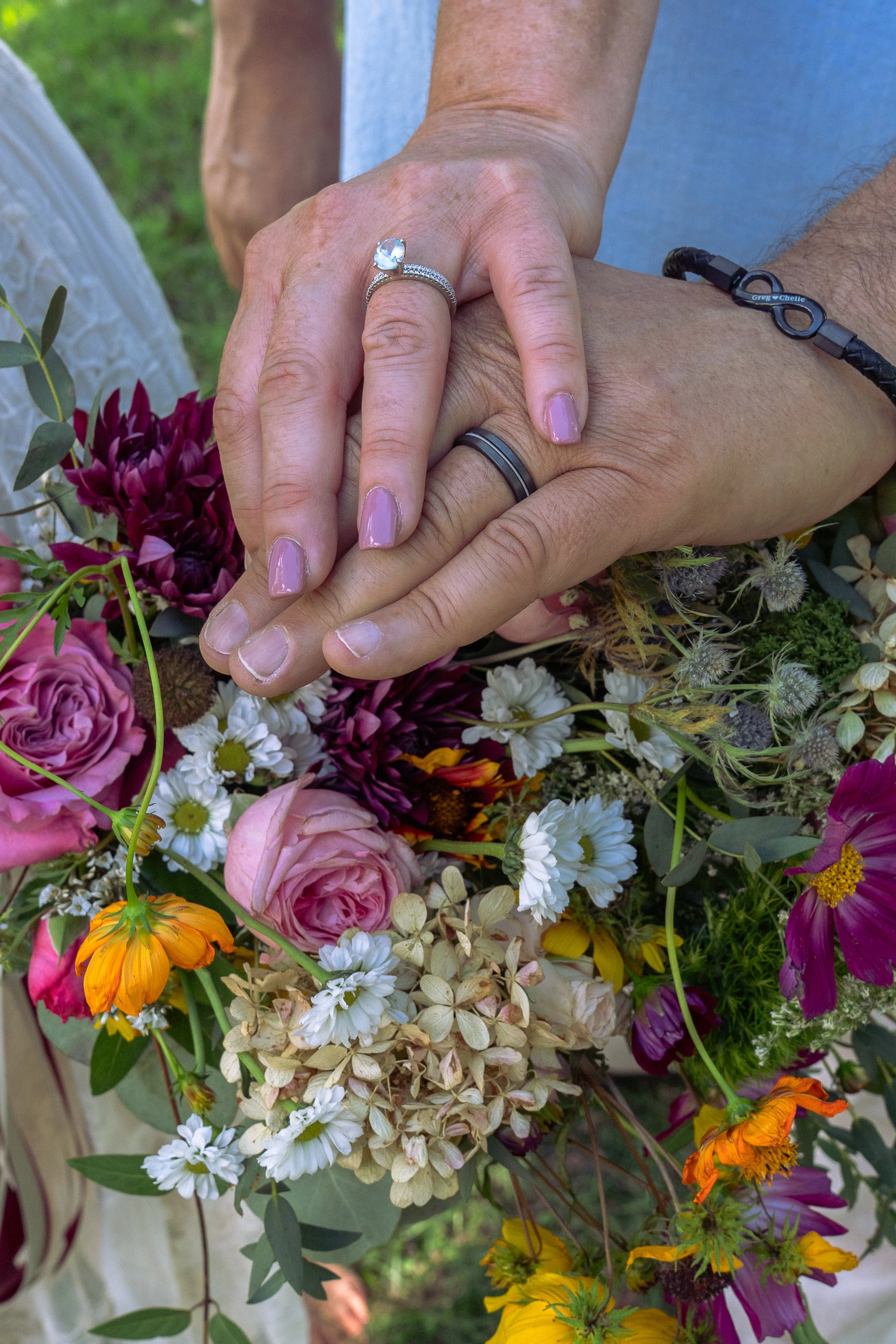 Close-up of two people's hands with wedding rings resting on top of a diverse bouquet of colorful flowers, including roses, daisies, and other blooms, set outdoors with greenery in the background.