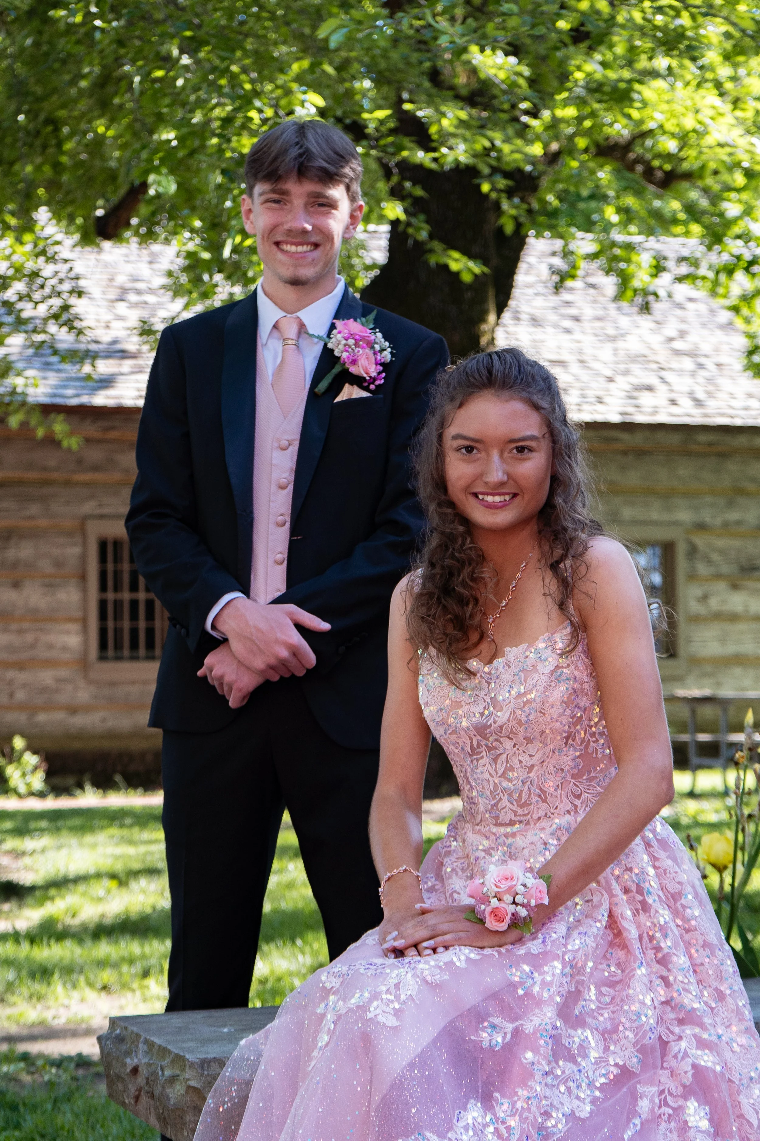 Young man in formal suit with a pink tie and boutonniere, and young woman in a pink lace gown with floral corsage, outdoors in front of a large tree, smiling for a photo.