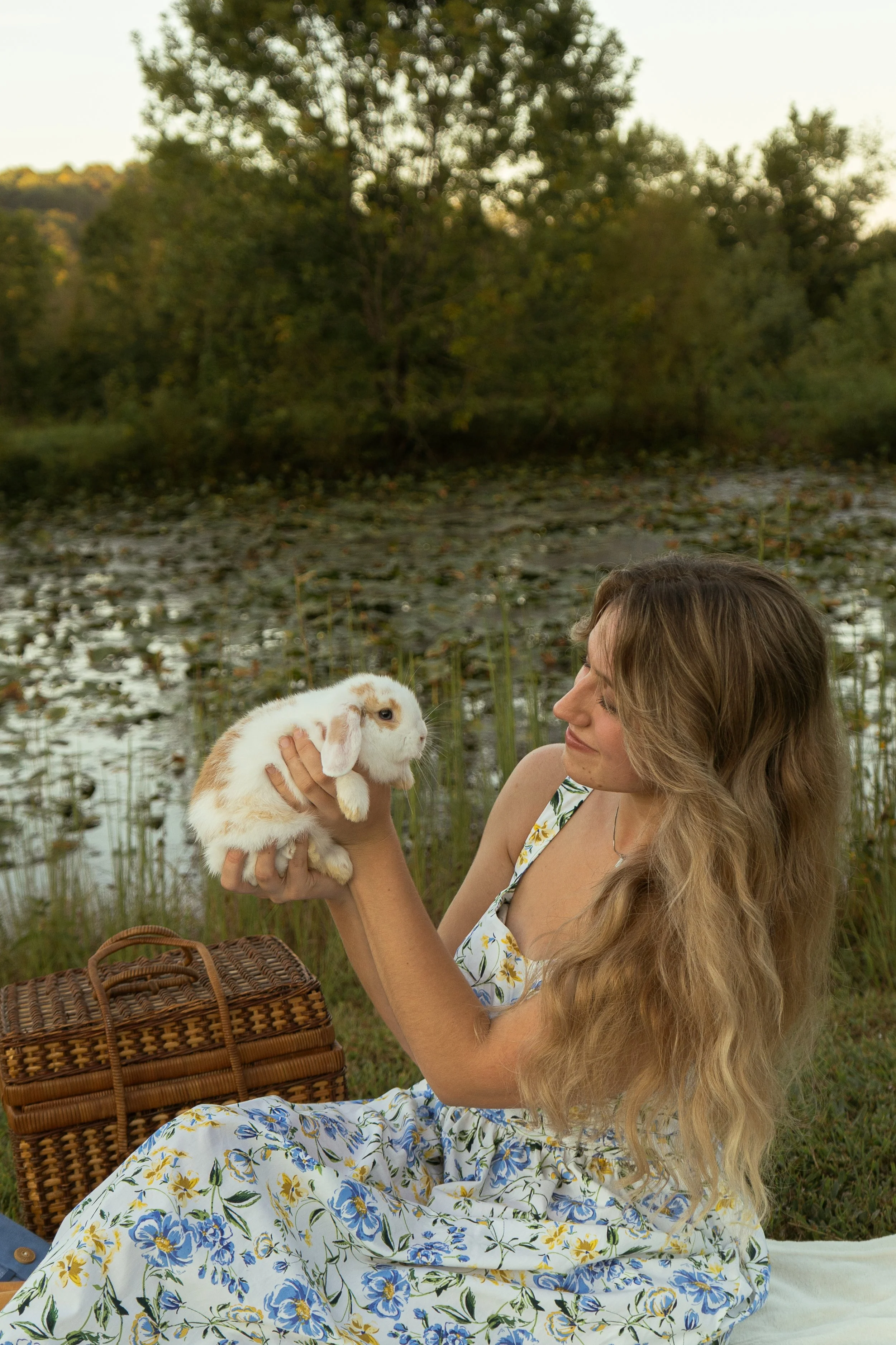 A woman with long wavy blonde hair, wearing a white dress with blue and yellow floral patterns, is sitting outdoors by a body of water surrounded by trees. She is holding a small white rabbit with light brown spots in her hands and looking at it. The