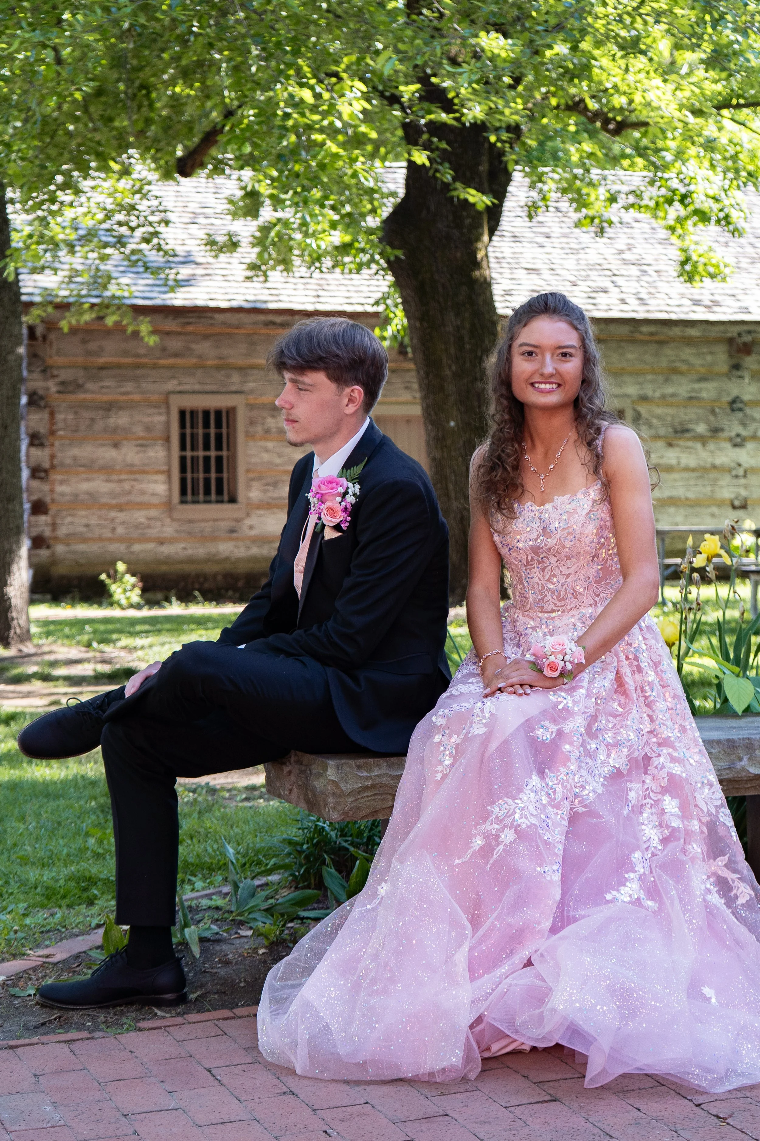 A young man in a dark suit with a pink boutonniere and a young woman in a pink lace gown with a floral bouquet sitting outdoors under a large tree on a sunny day, with a rustic wooden building in the background.