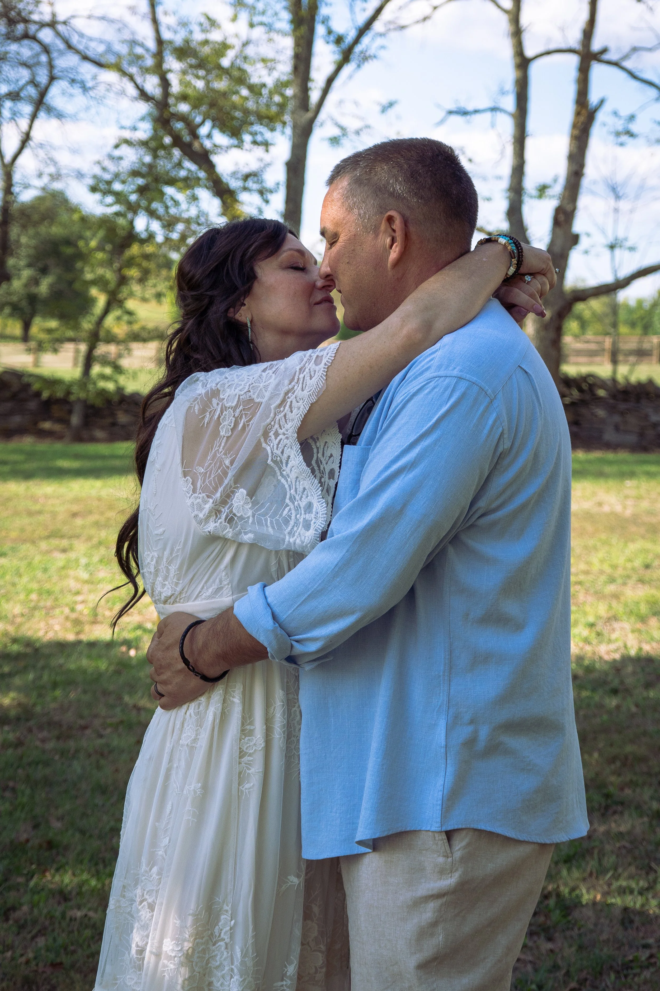 A woman in a white dress and a man in a blue shirt embracing and about to kiss outdoors with trees and a grassy area in the background.