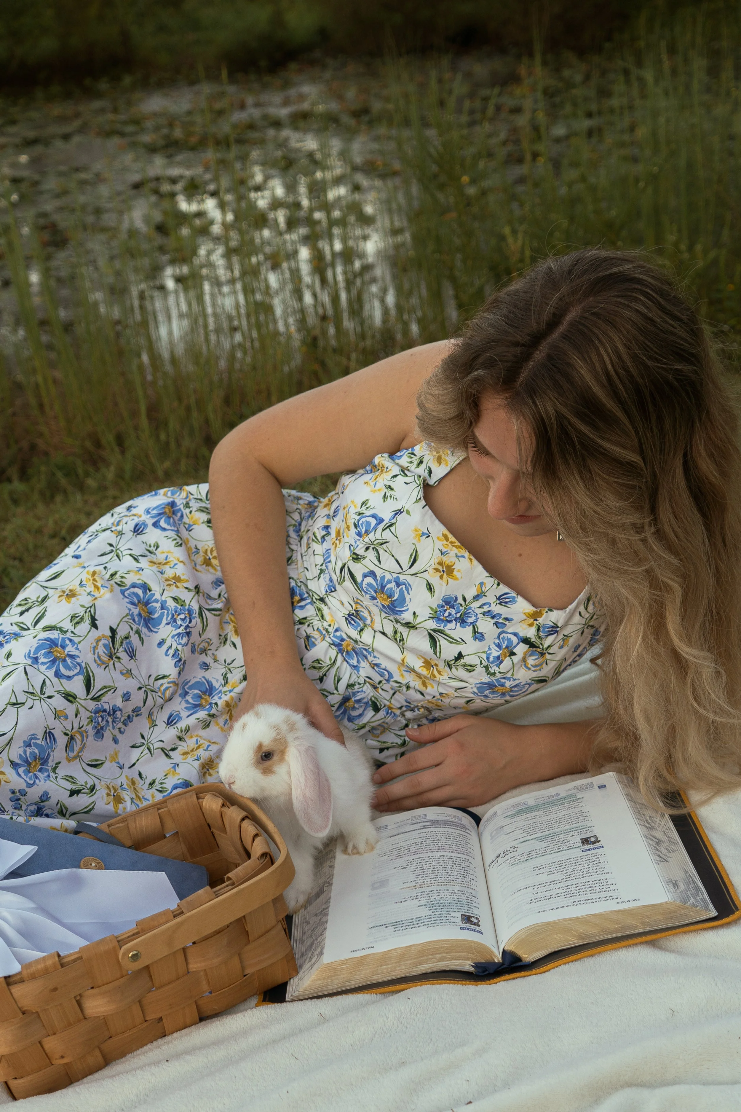 A woman with wavy brown hair wearing a floral dress relaxes outdoors by a pond with reeds. She is reading a book placed on a white blanket. A rabbit with white and brown fur and pink ears is near her, and a wicker basket filled with papers and a blue