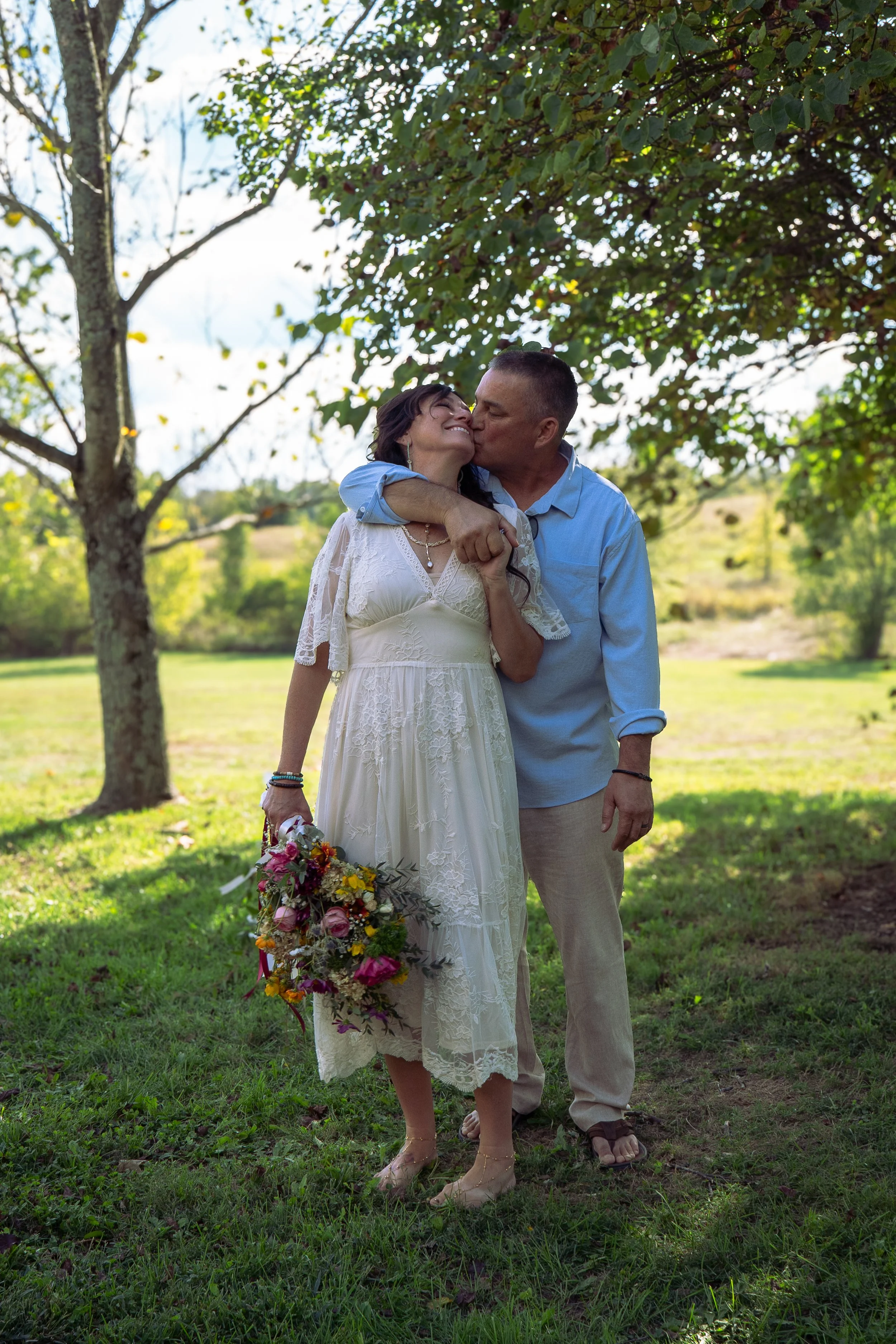 A couple dressed in summer clothing sharing a kiss outdoors, with the woman holding a large bouquet of colorful flowers, standing on grass with trees and a field in the background under a partly cloudy sky.