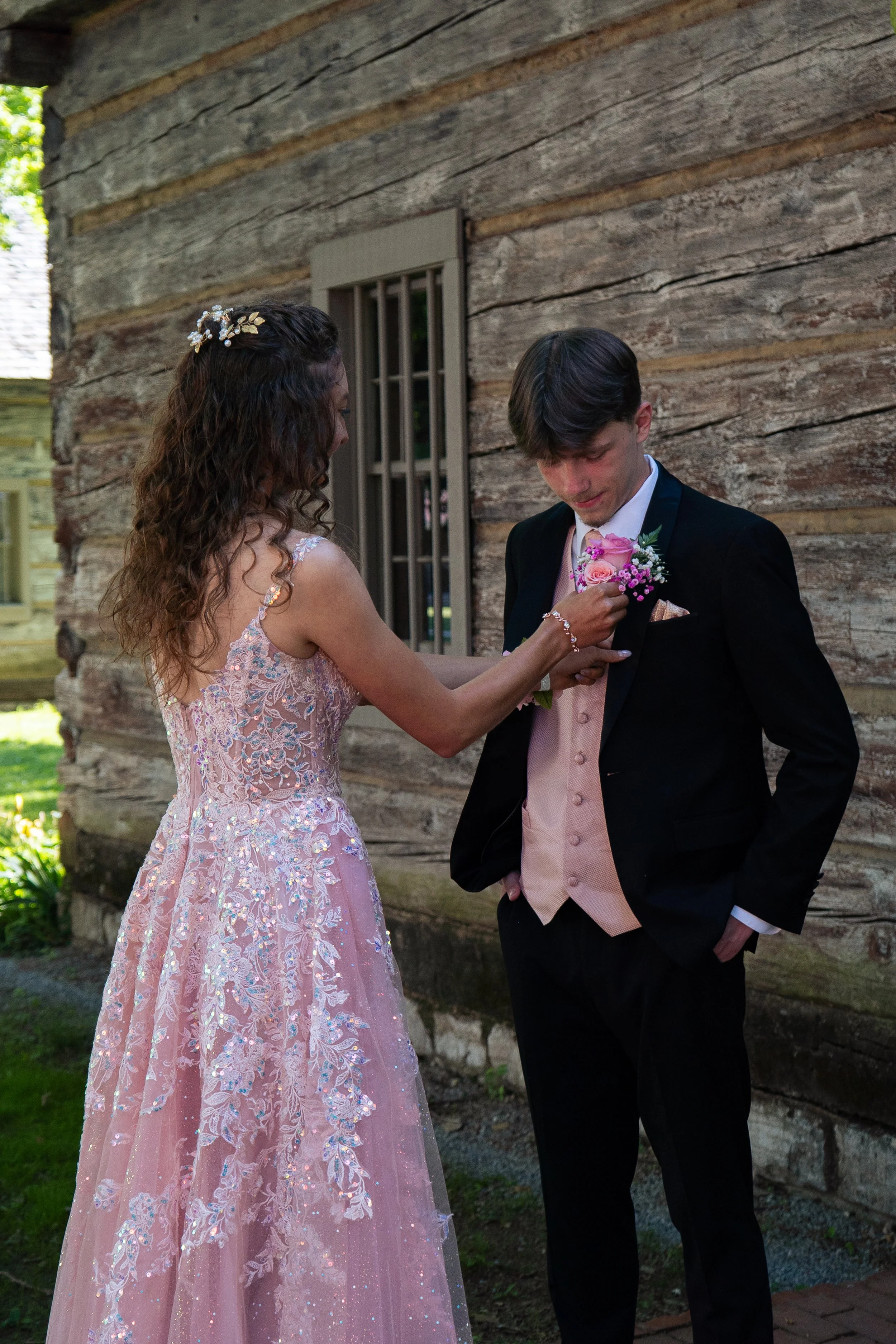 A woman in a pink, embroidered gown presenting a boutonniere to a man in a black tuxedo with a pink vest and pink boutonniere, standing outdoors next to a rustic wooden wall.