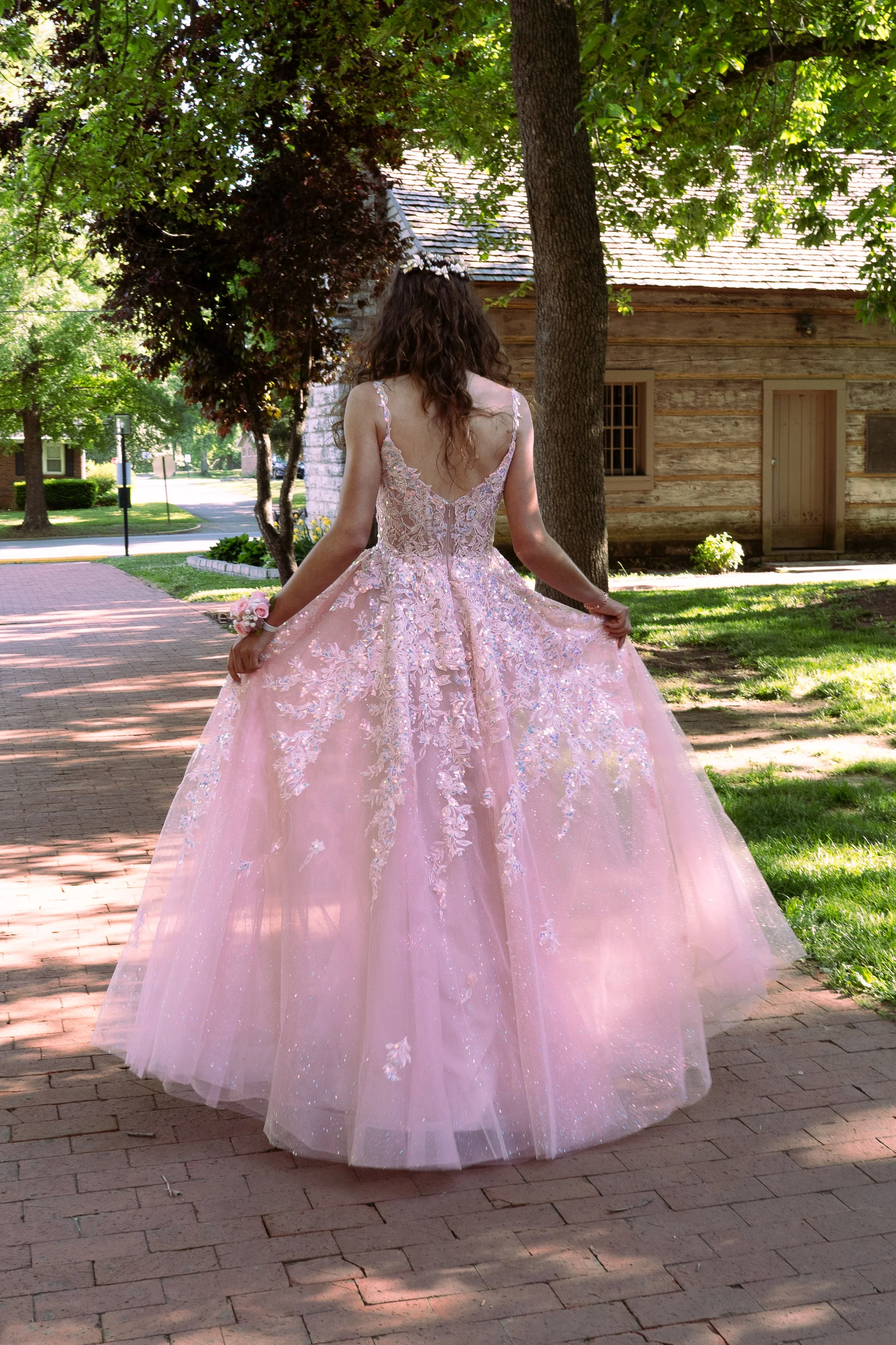 A woman in a pink ball gown with lace and embroidery details, standing outdoors on a brick pathway under trees, with a rustic wooden building in the background.