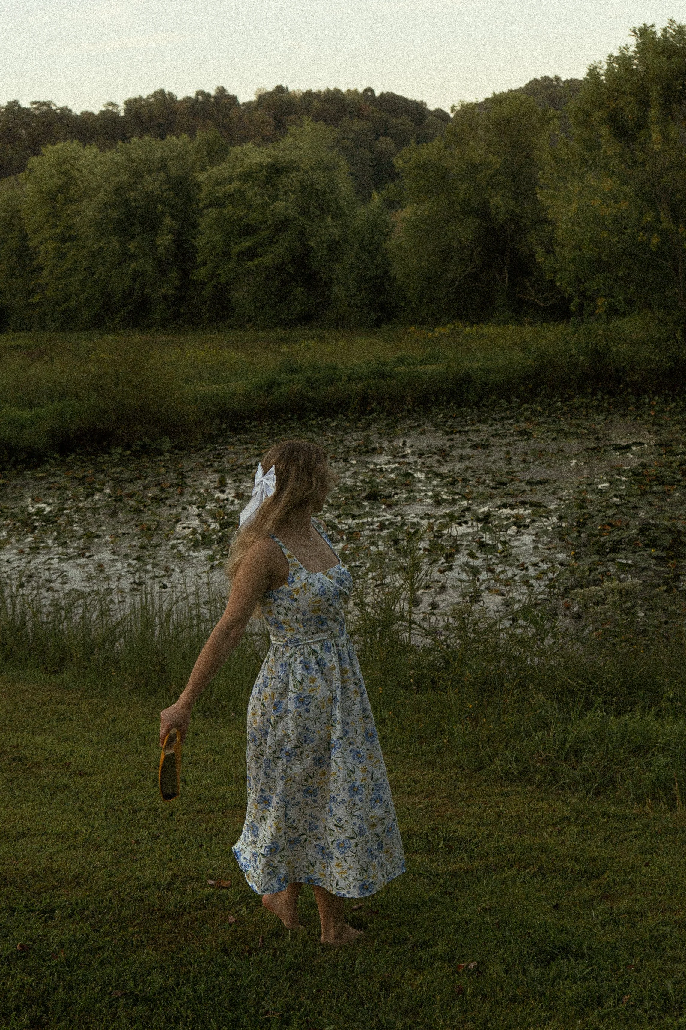A woman with long hair wearing a floral dress and a white bow in her hair walking barefoot on grass near a wetland area with trees and bushes in the background.
