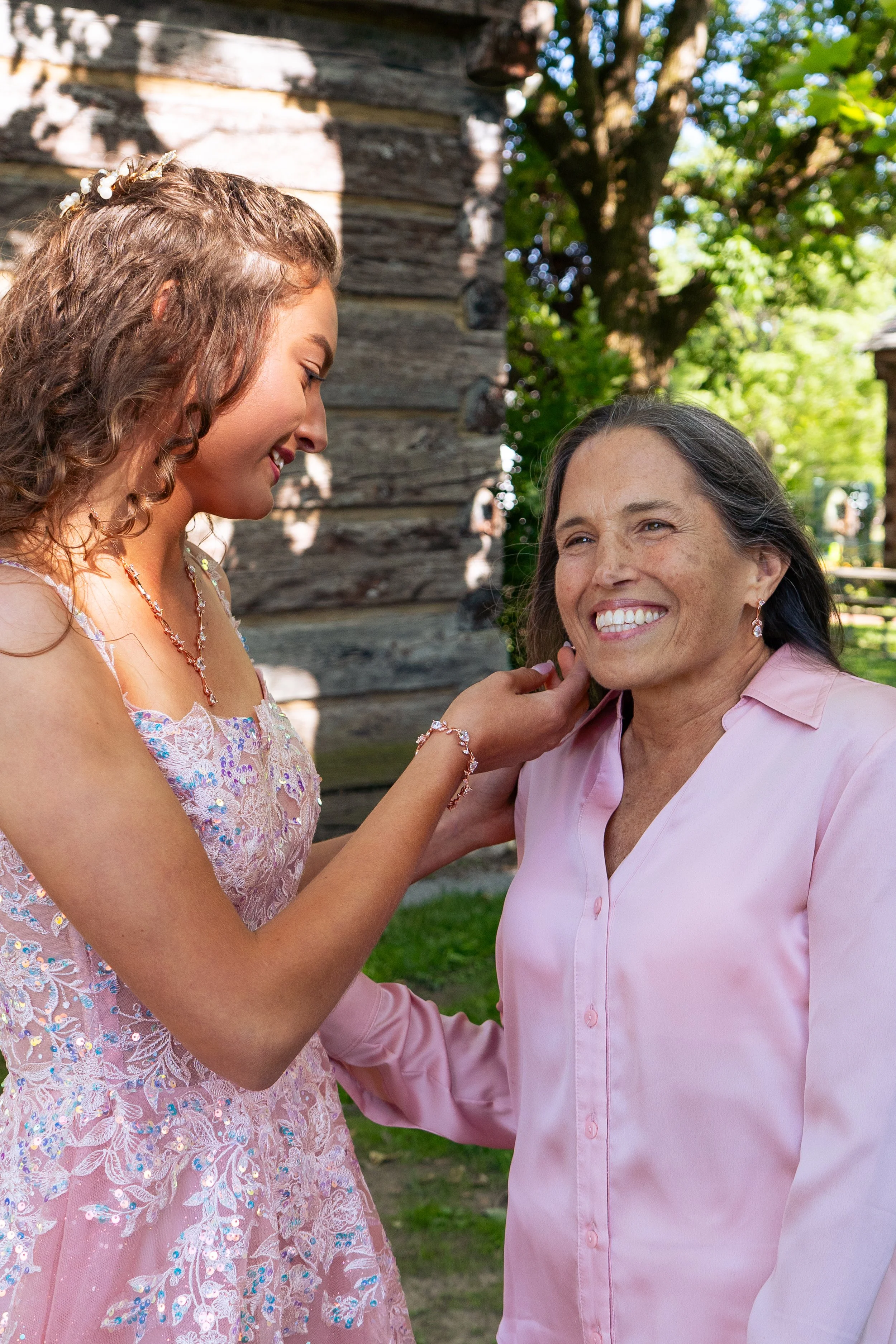 A young woman in a pink dress and floral jewelry touches the face of an older woman with gray hair and a pink shirt, both smiling, outdoors near a tree and a rustic wooden building.