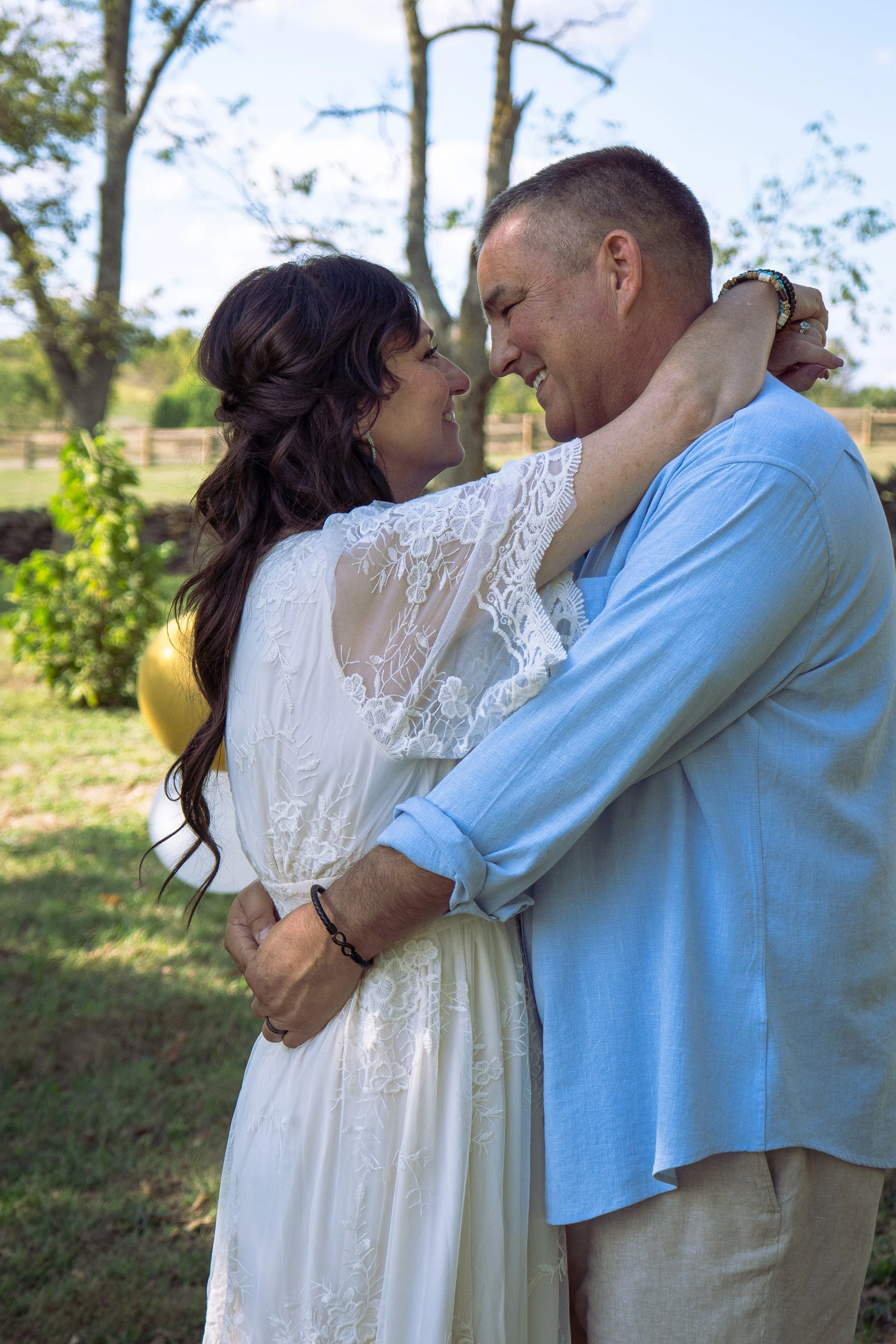 A couple embraces outdoors, smiling at each other, with trees and a fence in the background on a sunny day.
